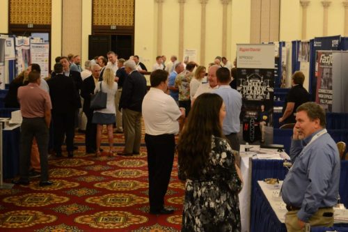 Busy conference expo hall with people mingling among booth displays and banners along blue-curtained booths.