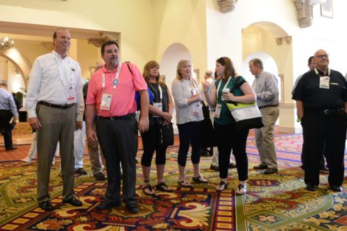 Group of conference attendees standing in a lobby with arched columns and a colorful patterned carpet, wearing lanyards and badges.