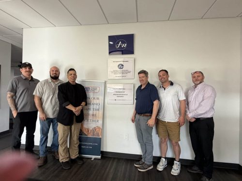 Seven men stand in a line in a corporate lobby, posing for a photo by wall-mounted signs and plaques.