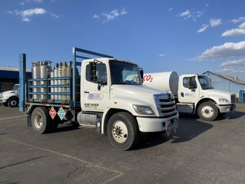 Flatbed trailer loaded with tall gas cylinders next to a white delivery truck at a welding supply yard in an outdoor lot.