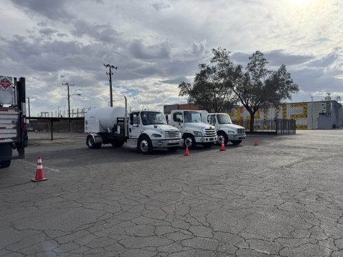 Row of white commercial trucks parked in a lot under a cloudy sky, with orange safety cones in the foreground.