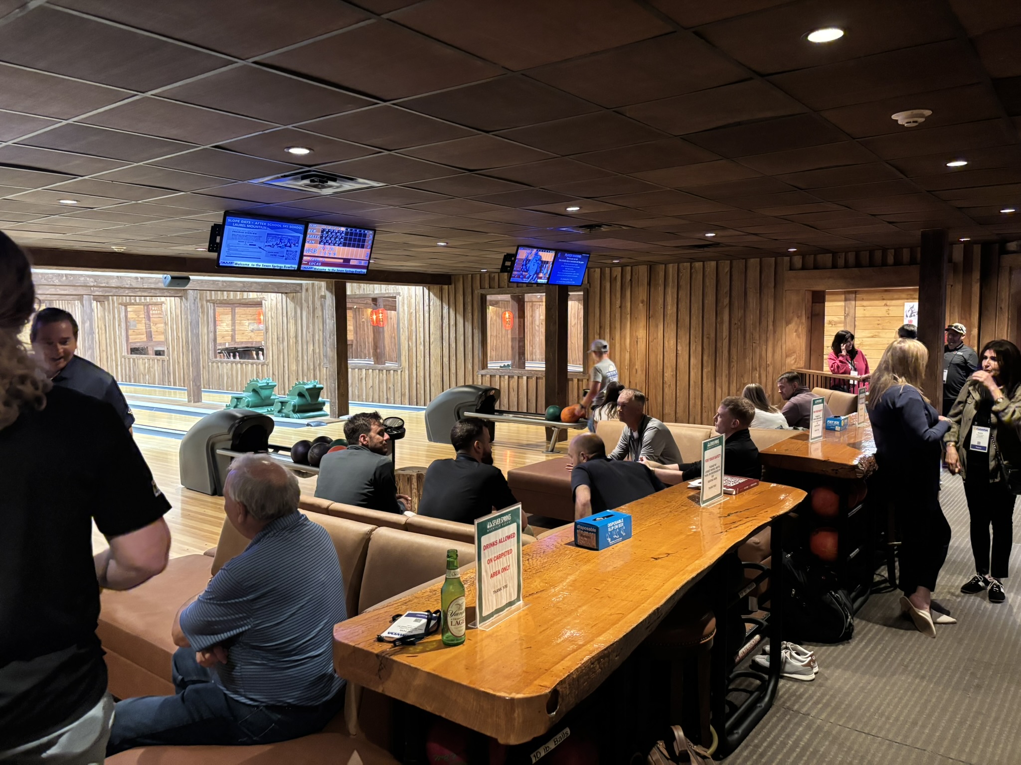 Indoor bowling alley with lanes, seating tables, and people socializing under a brown tiled ceiling.