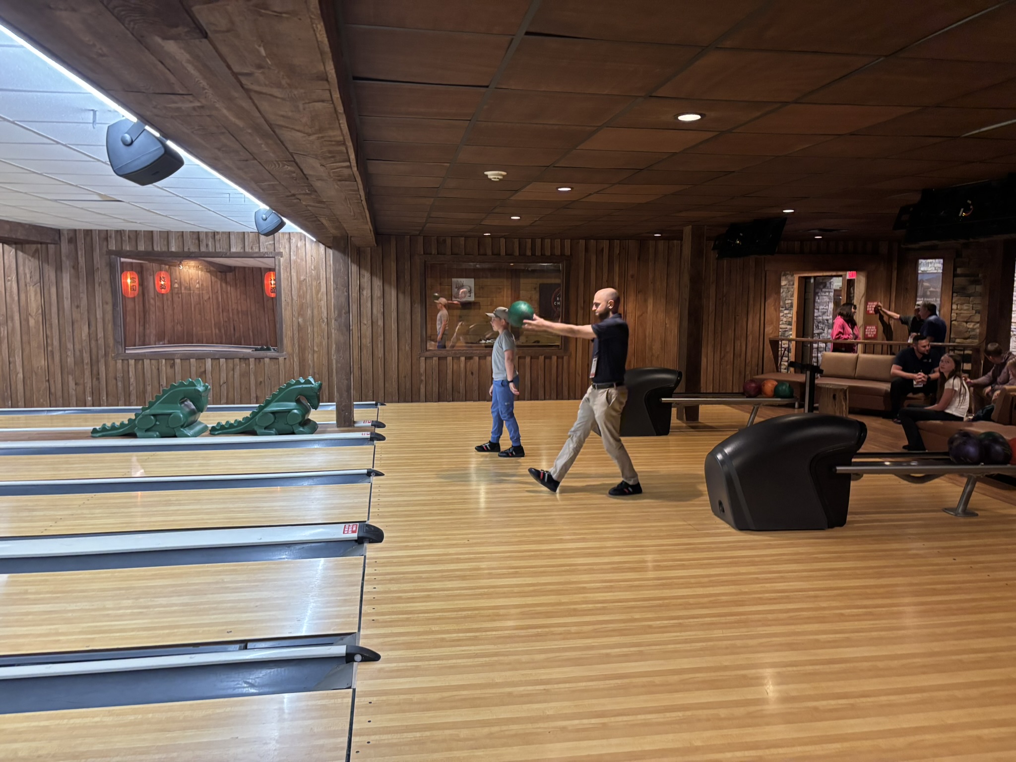 A bowling alley scene with wooden walls and ceiling, several lanes, green dragon bumper guards, and a man throwing a bowling ball while another person watches nearby.