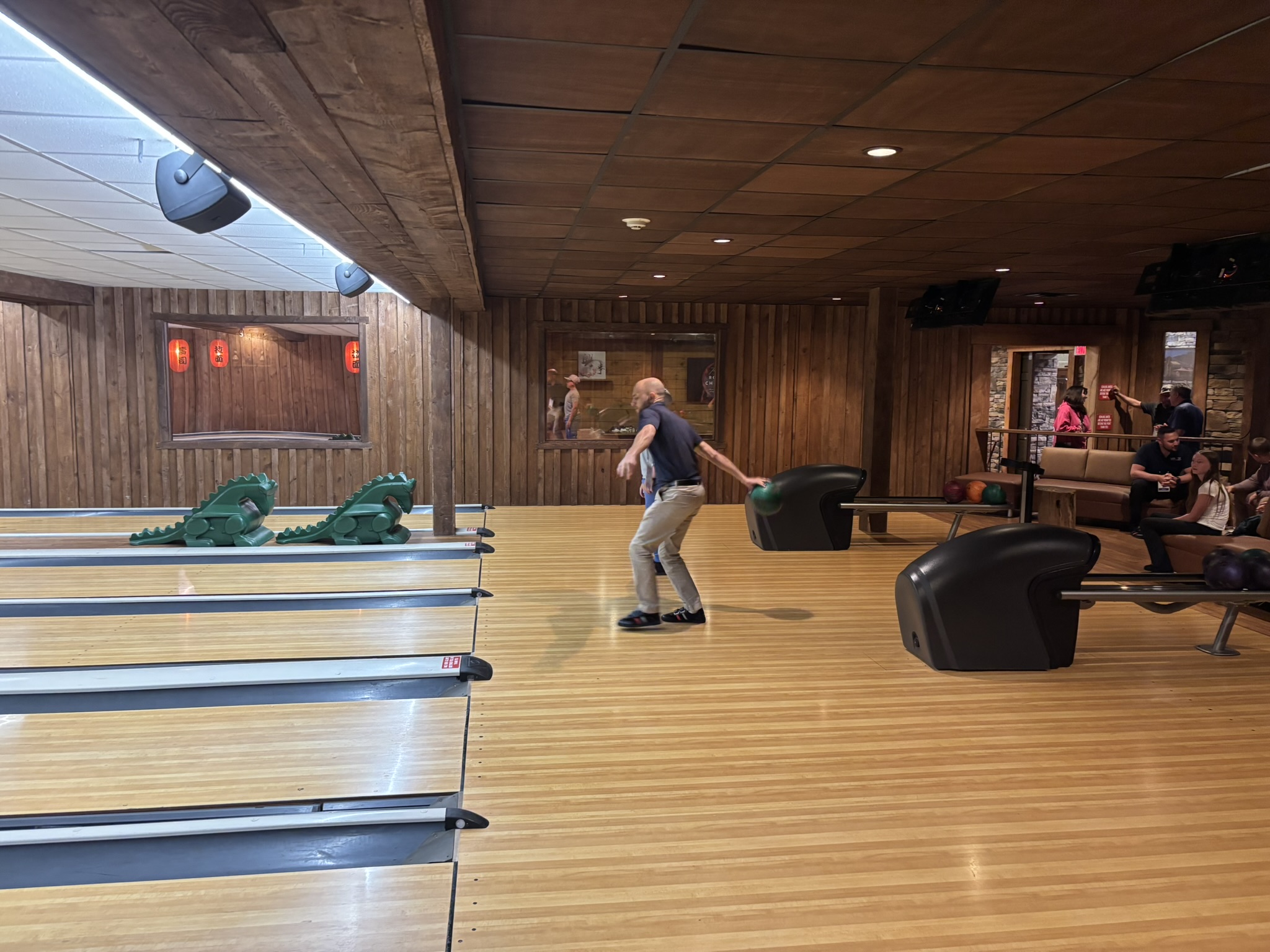 A man in a dark shirt bowls on a wooden bowling lane, with spectators sitting in a lounge area to the right and ball return machines nearby.