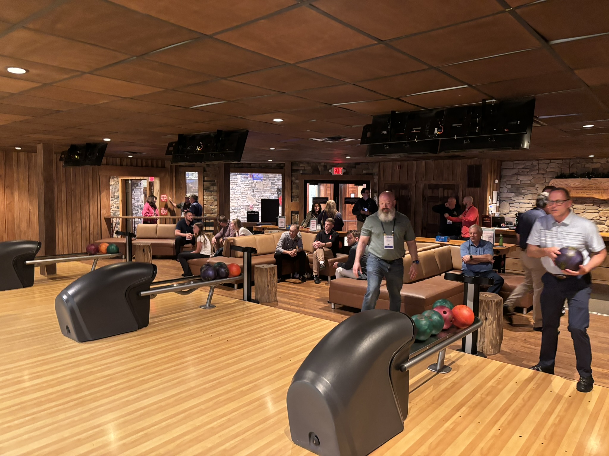 People gather in a bowling alley with lanes, seating area, and ball racks in the foreground; players wait and chat as others bowl.