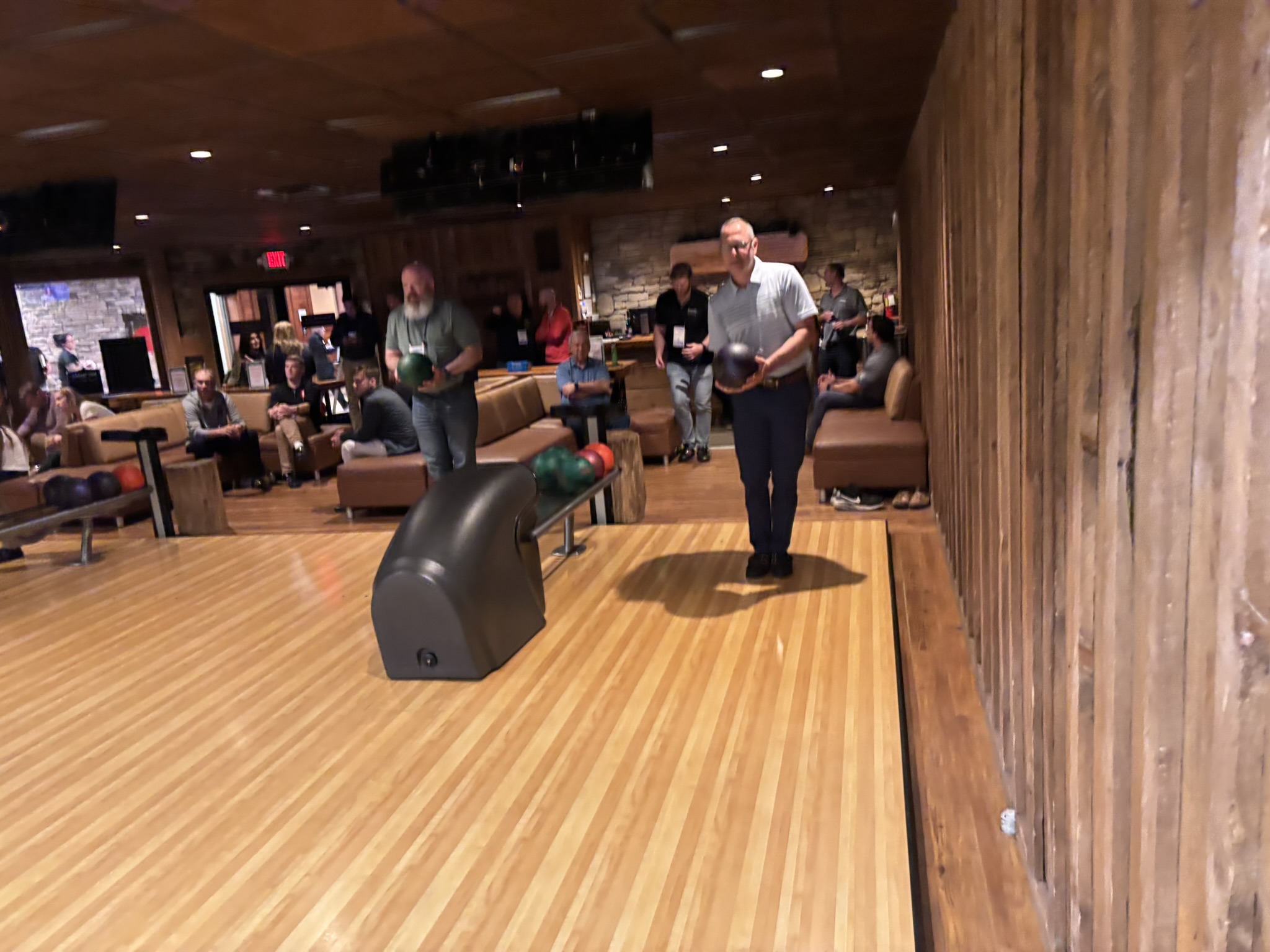 Bowling alley scene with people gathered around lanes; a man in a light shirt stands at the lane holding a bowling ball while others watch in the background.