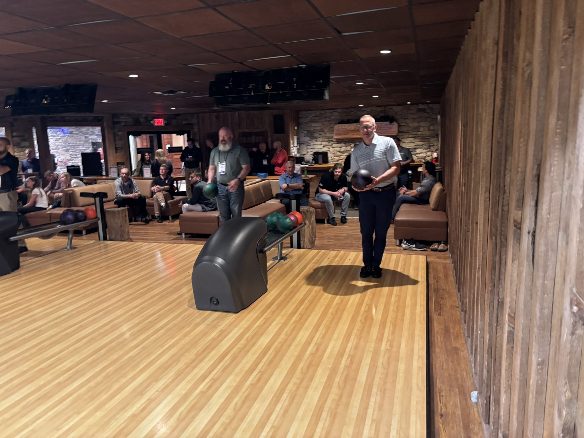Two men stand at the end of a bowling lane with balls in hand while a group watches from behind on couches in a wood-paneled bowling alley.