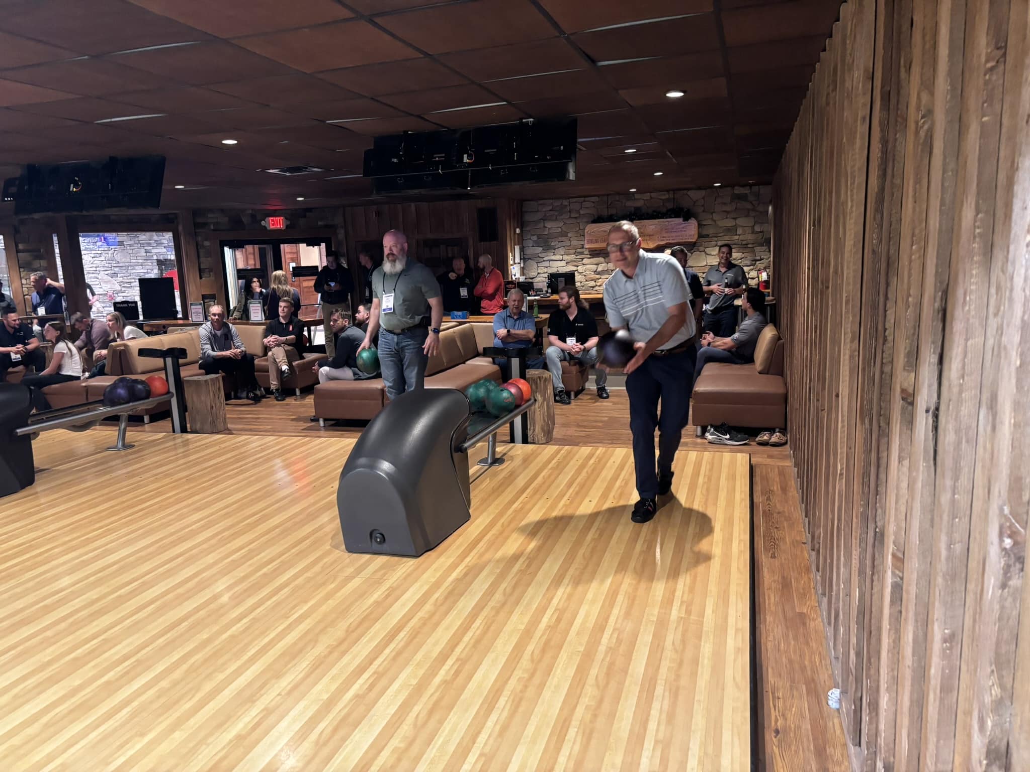 Man mid-throw at a bowling lane, with spectators seated and watching in a wood-paneled alley.