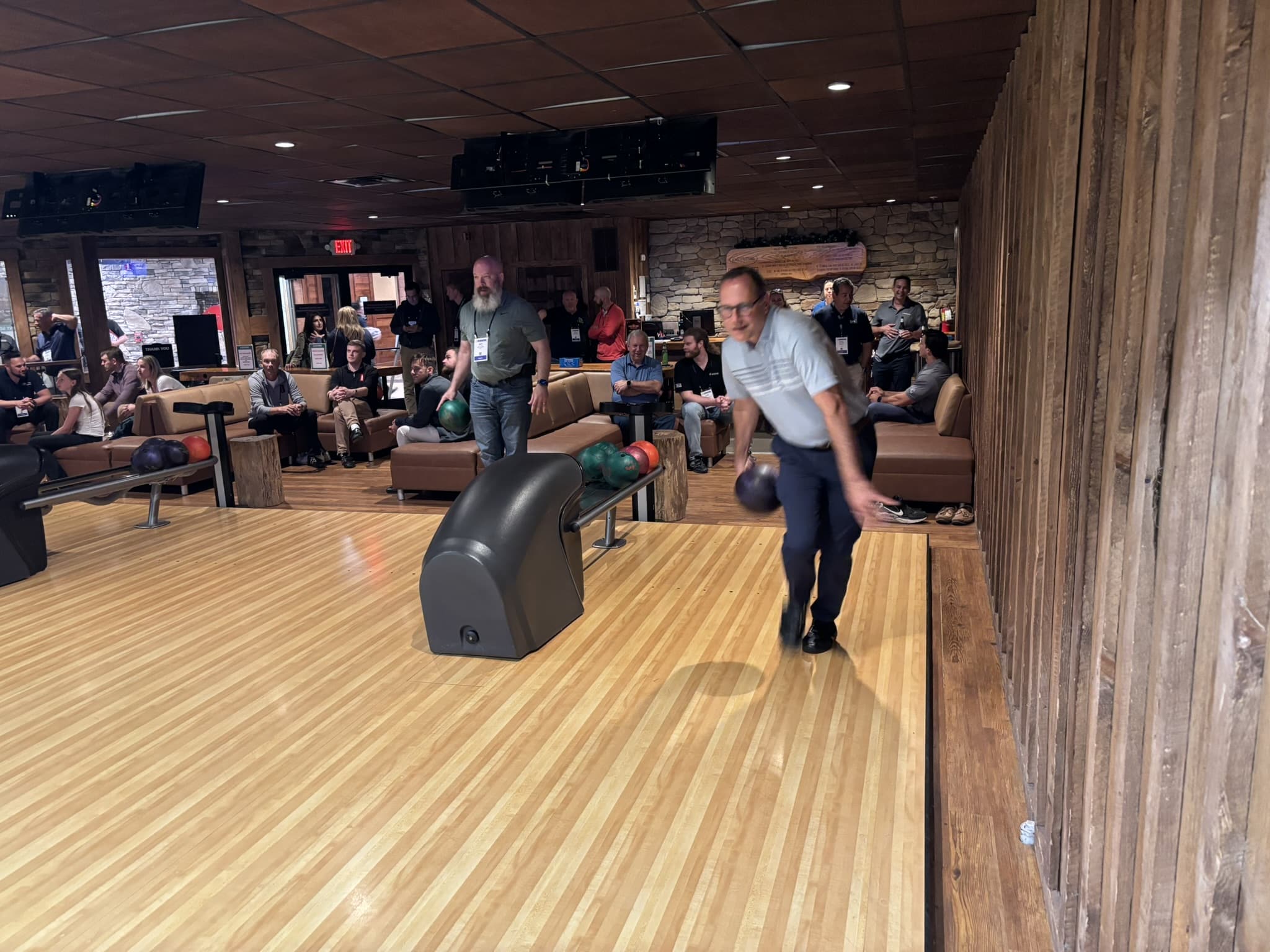 People seated along bowling lanes watching a game; a bowler in a light blue shirt is mid-throw in the foreground.