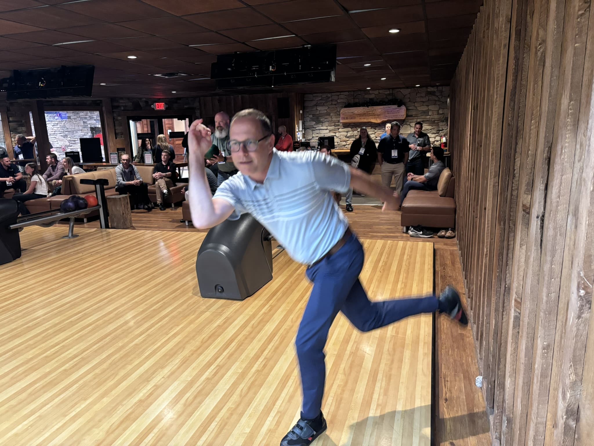Bulking bowler in a light blue shirt throwing a ball down a wooden bowling lane while spectators sit and watch in the background.