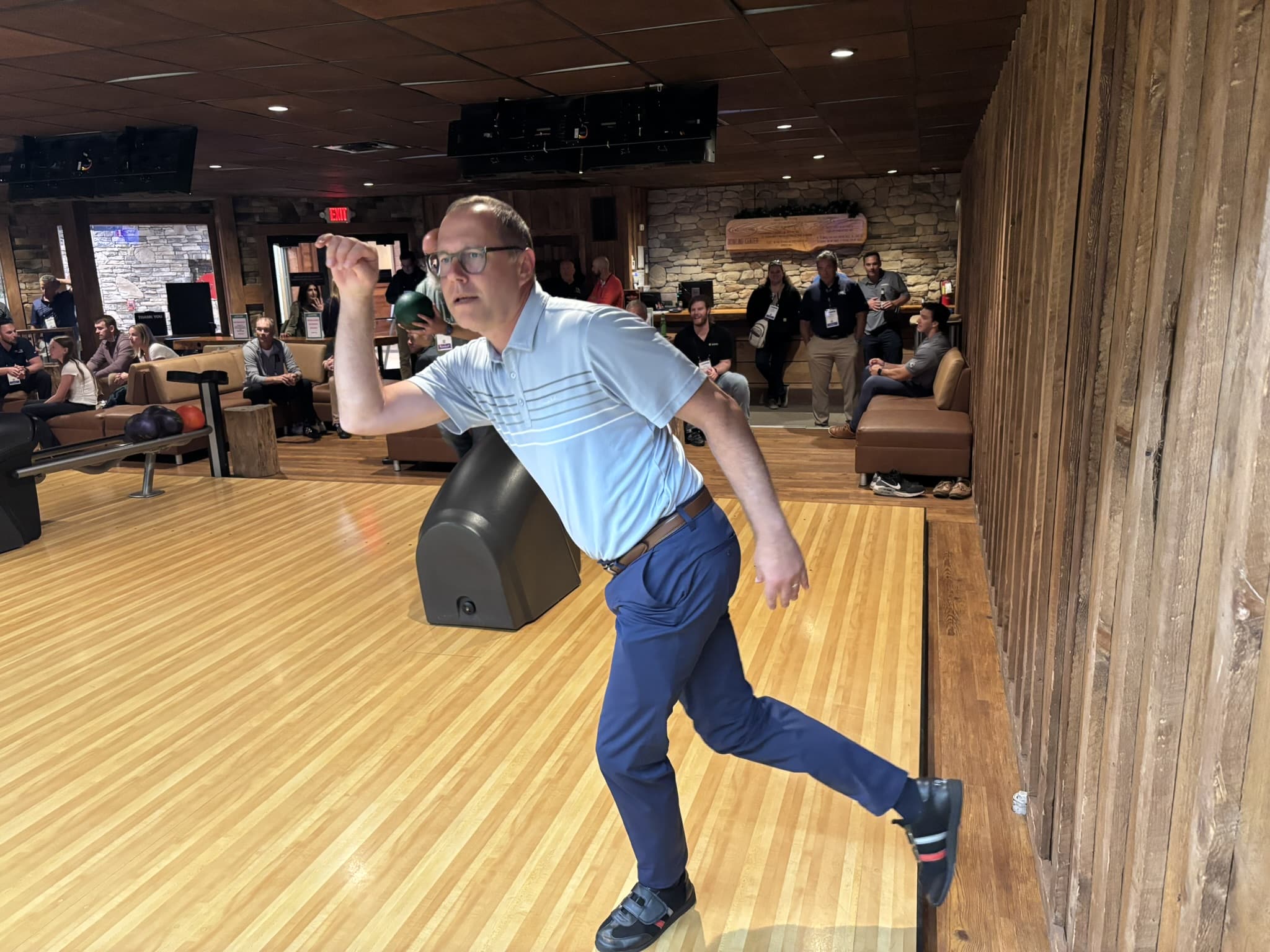 Man in a light blue polo and blue pants mid-throw in a bowling alley, with onlookers seated and standing in the background.