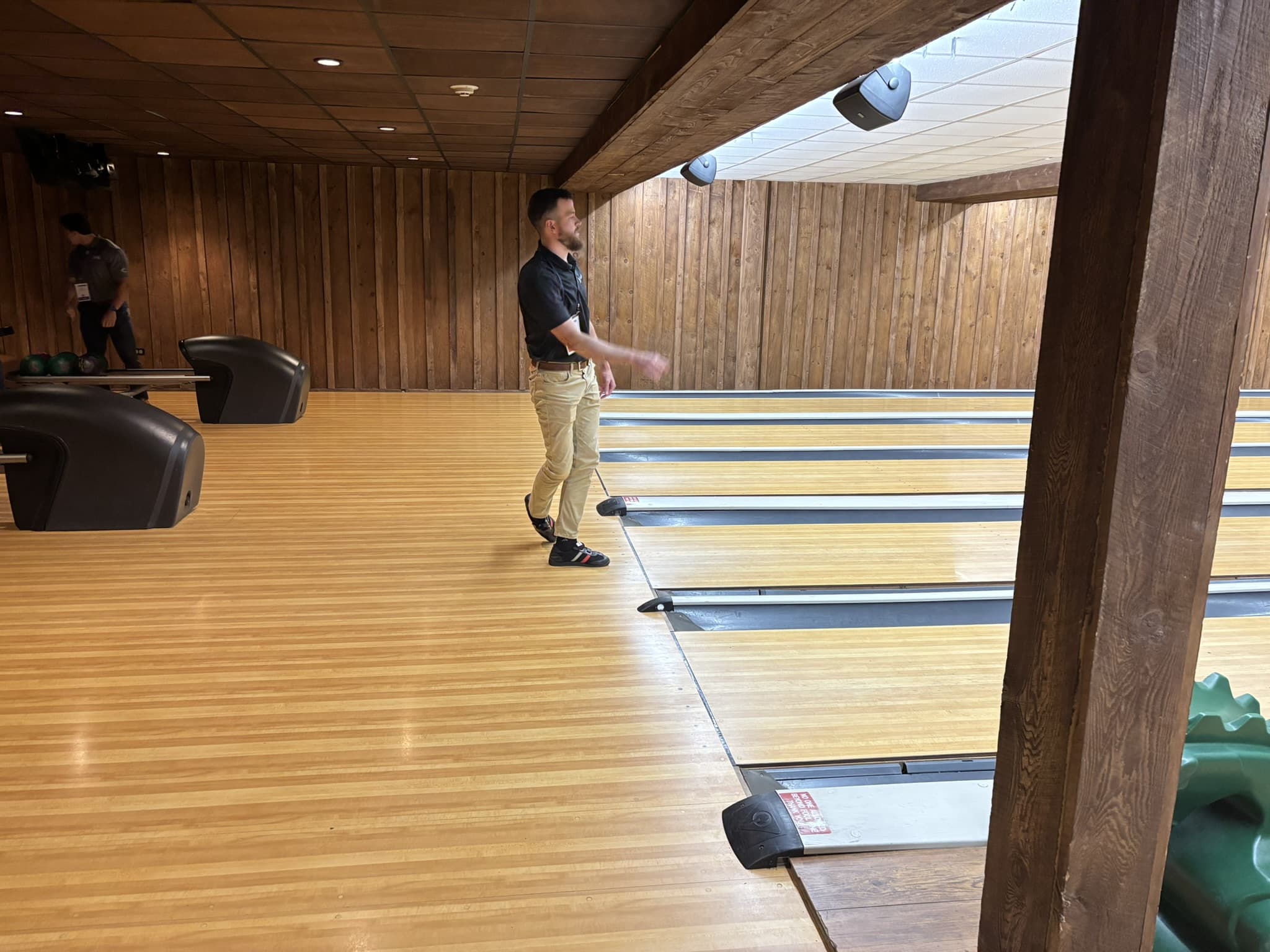 Man in a dark polo and khaki pants bowling on a wooden lane inside a wood-paneled bowling alley, with another person near the ball racks in the background.