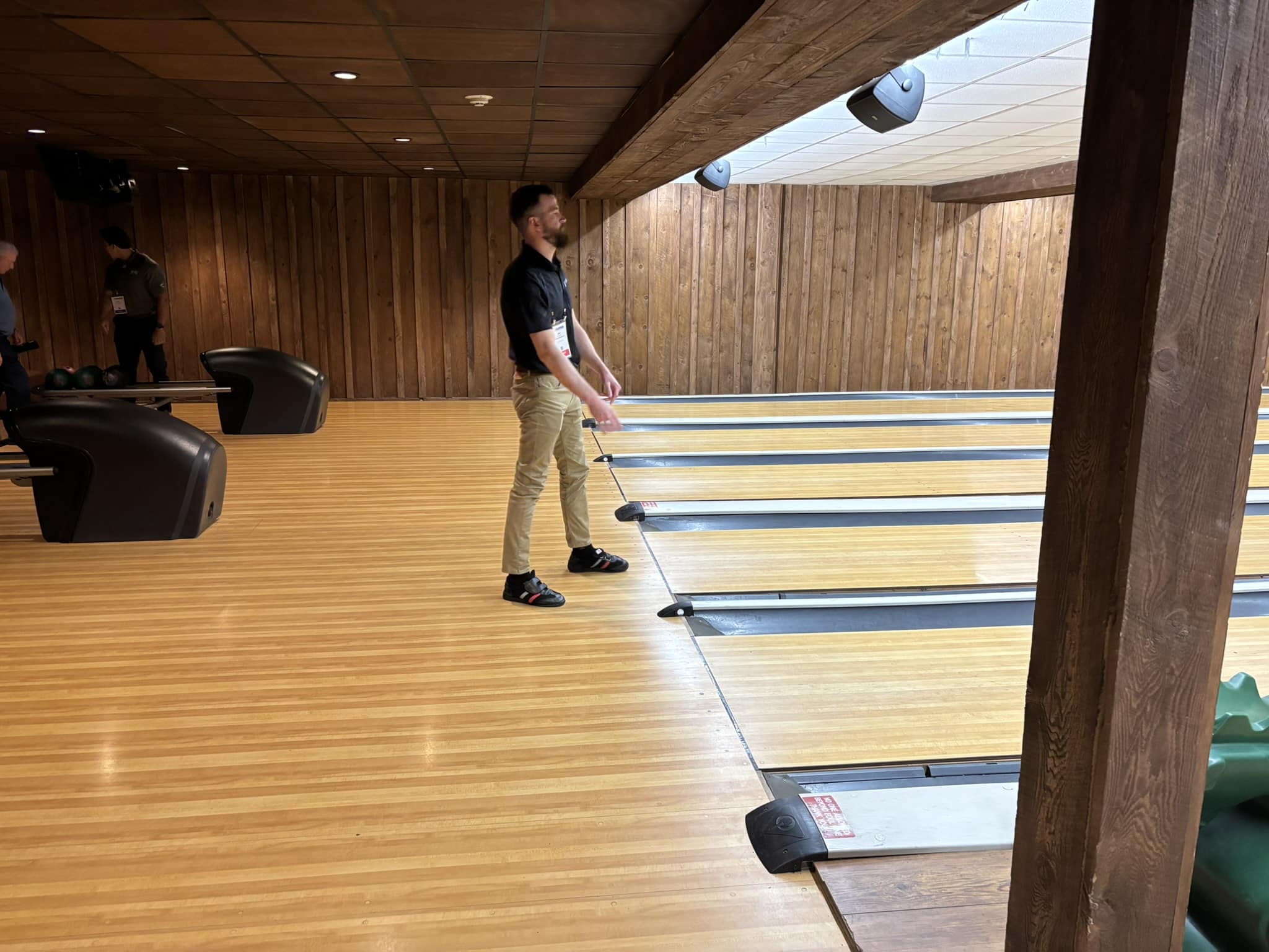 Man with a beard in a black shirt and khaki pants standing on a bowling lane, preparing to bowl in a wooden bowling alley with others in the background.