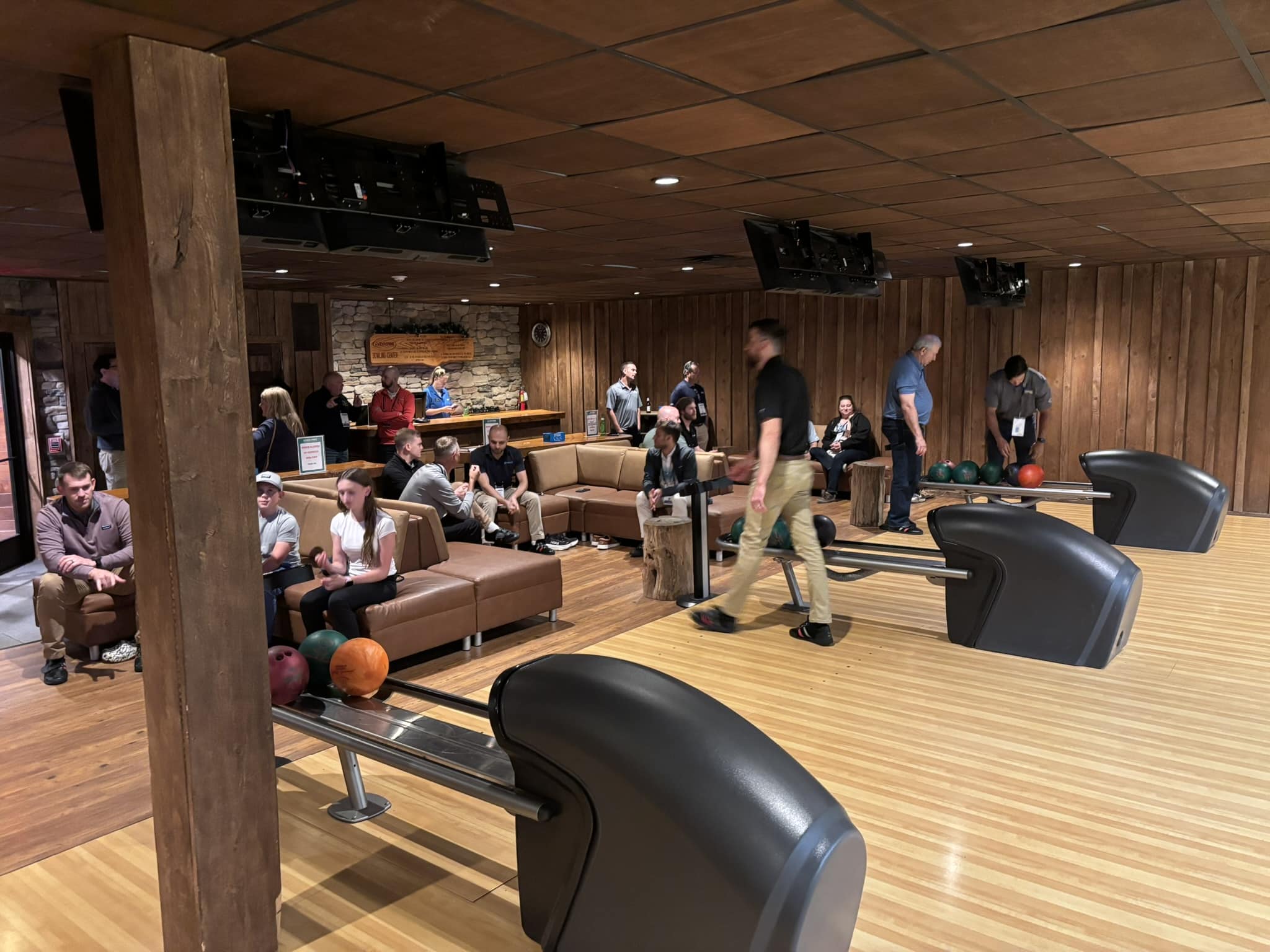 Bowling alley interior with wooden walls and ceiling, people sitting on brown couches, and bowling balls on a rack near the lanes.