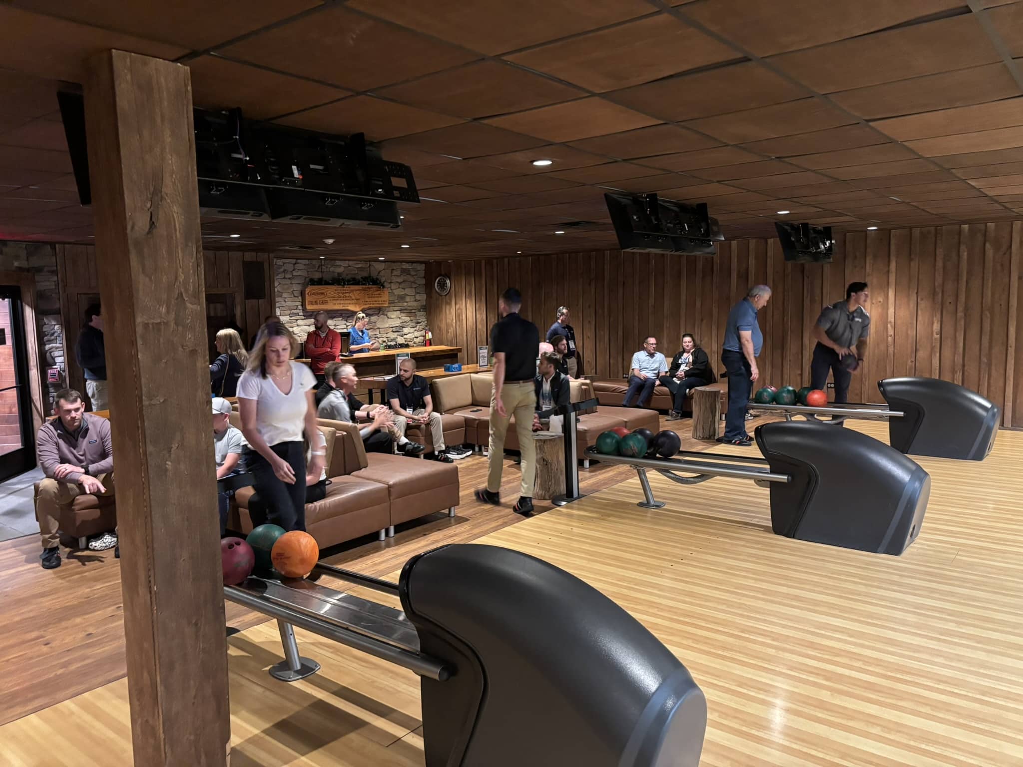 Bowling alley interior with wooden beams and lanes; people seated on couches and standing, watching or chatting near the ball racks and lane area.