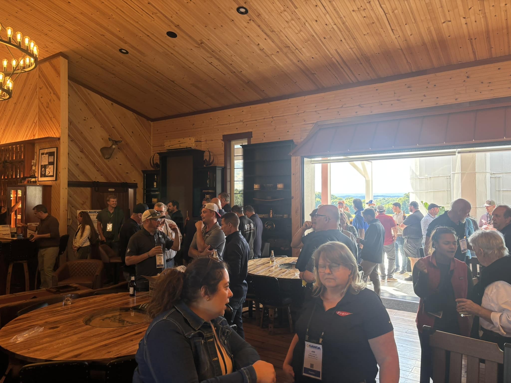 Crowd of attendees mingling at a rustic wood-paneled indoor event, many with name badges and drinks in hand.