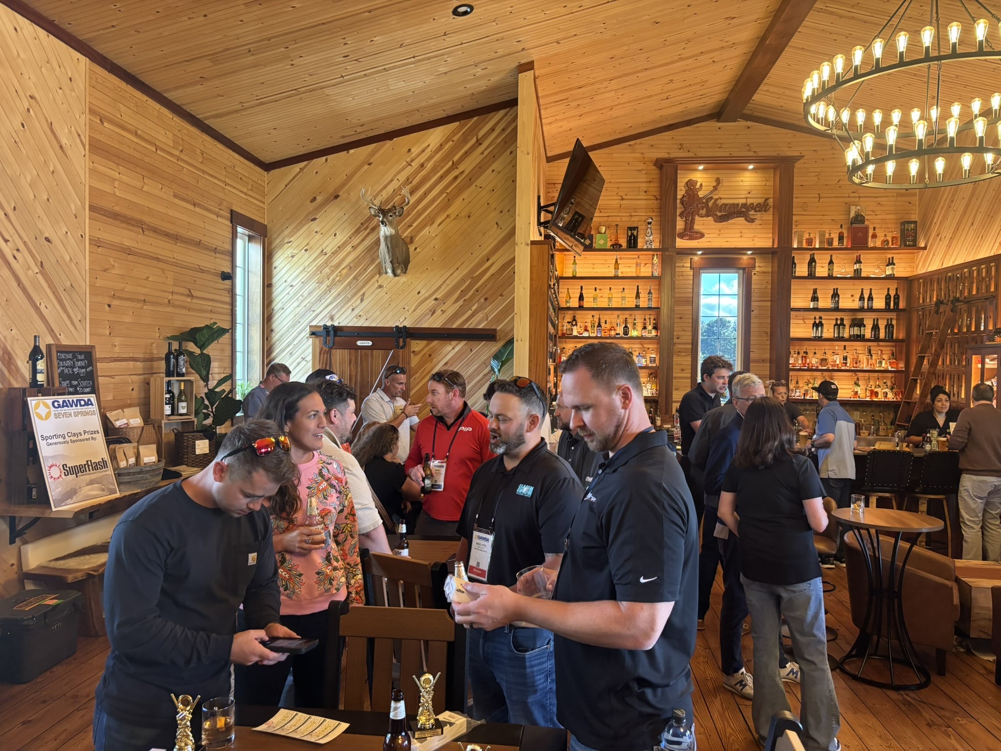 Crowd of people socializing at a rustic wooden bar, with liquor shelves, warm lighting, a circular chandelier, and a mounted deer head on the wall.