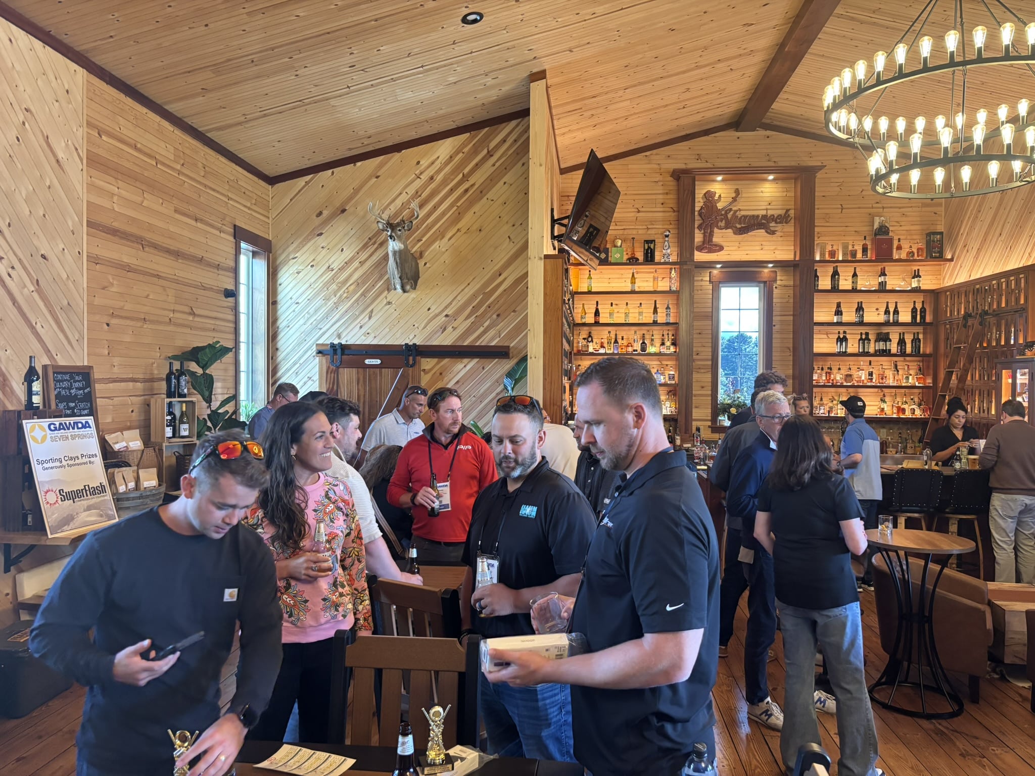 Crowded wooden-bar interior with shelves of bottles, a deer head mount, and a large chandelier overhead.