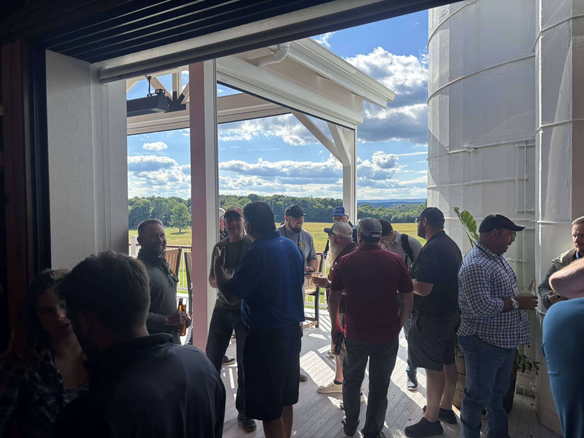 Group of people chatting and holding drinks on a covered outdoor deck, with a scenic field and blue sky in the background.