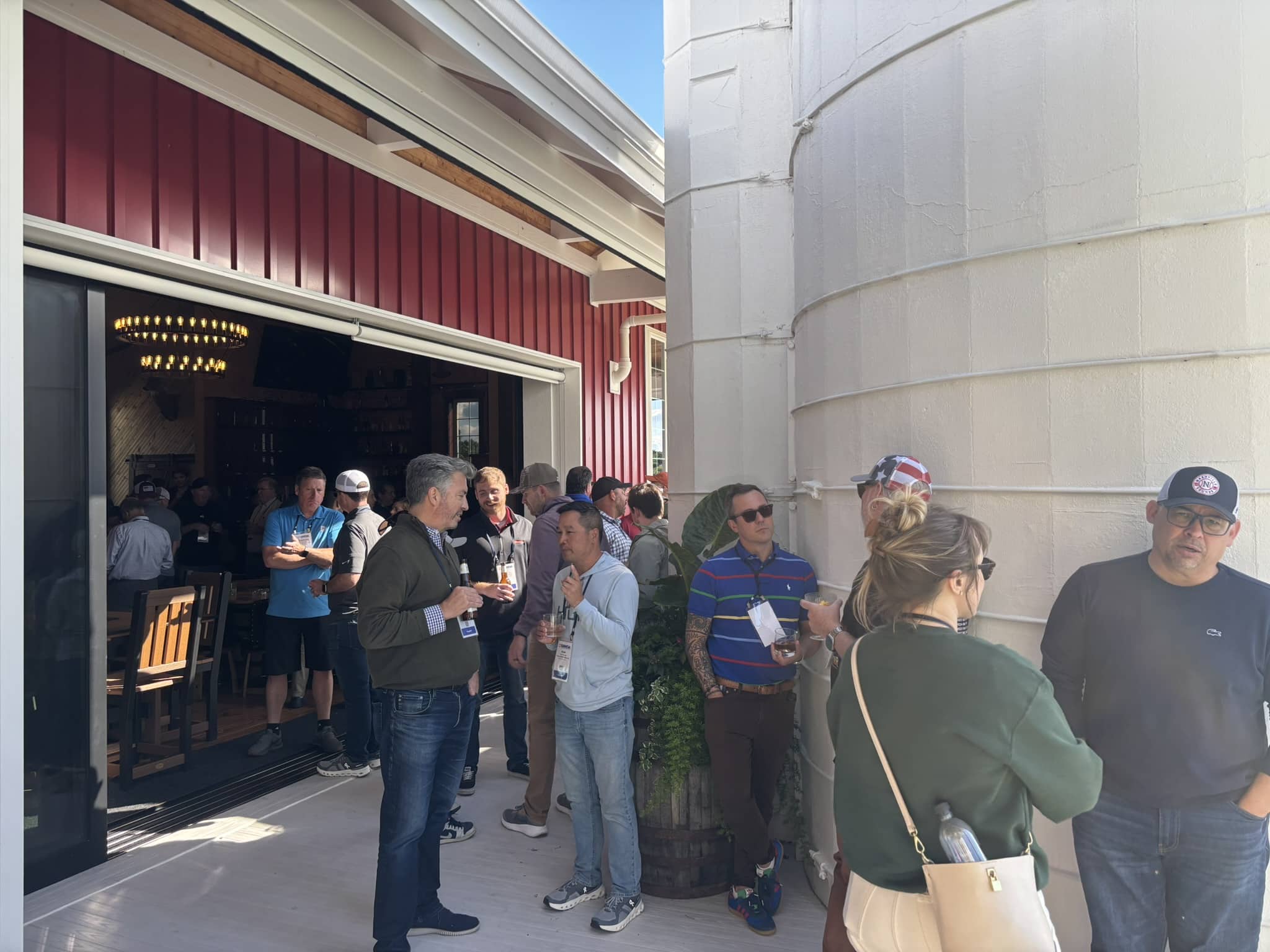 Crowd of people standing in line outside a red building, some holding drinks and wearing lanyards at a social event.