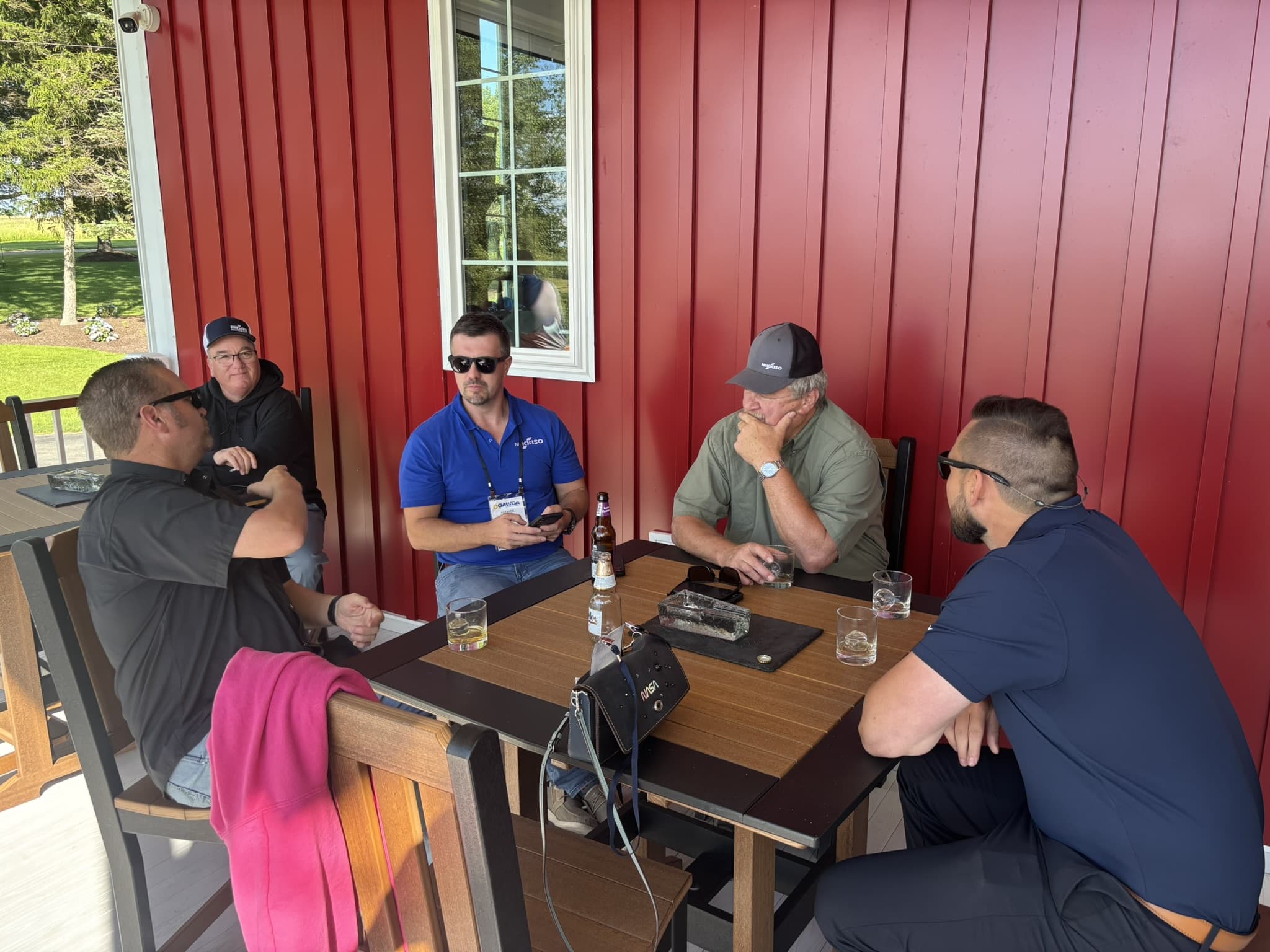 Five men sit around a wooden table on a covered patio with red vertical siding, chatting and having drinks.