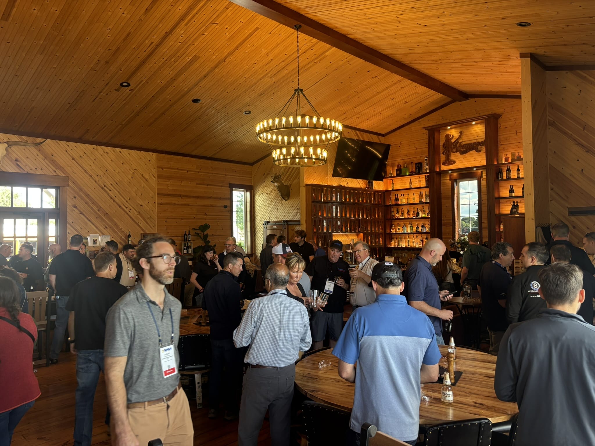 Busy, wood-paneled bar area with a circular chandelier and many guests wearing name badges mingling around tables and liquor shelves.