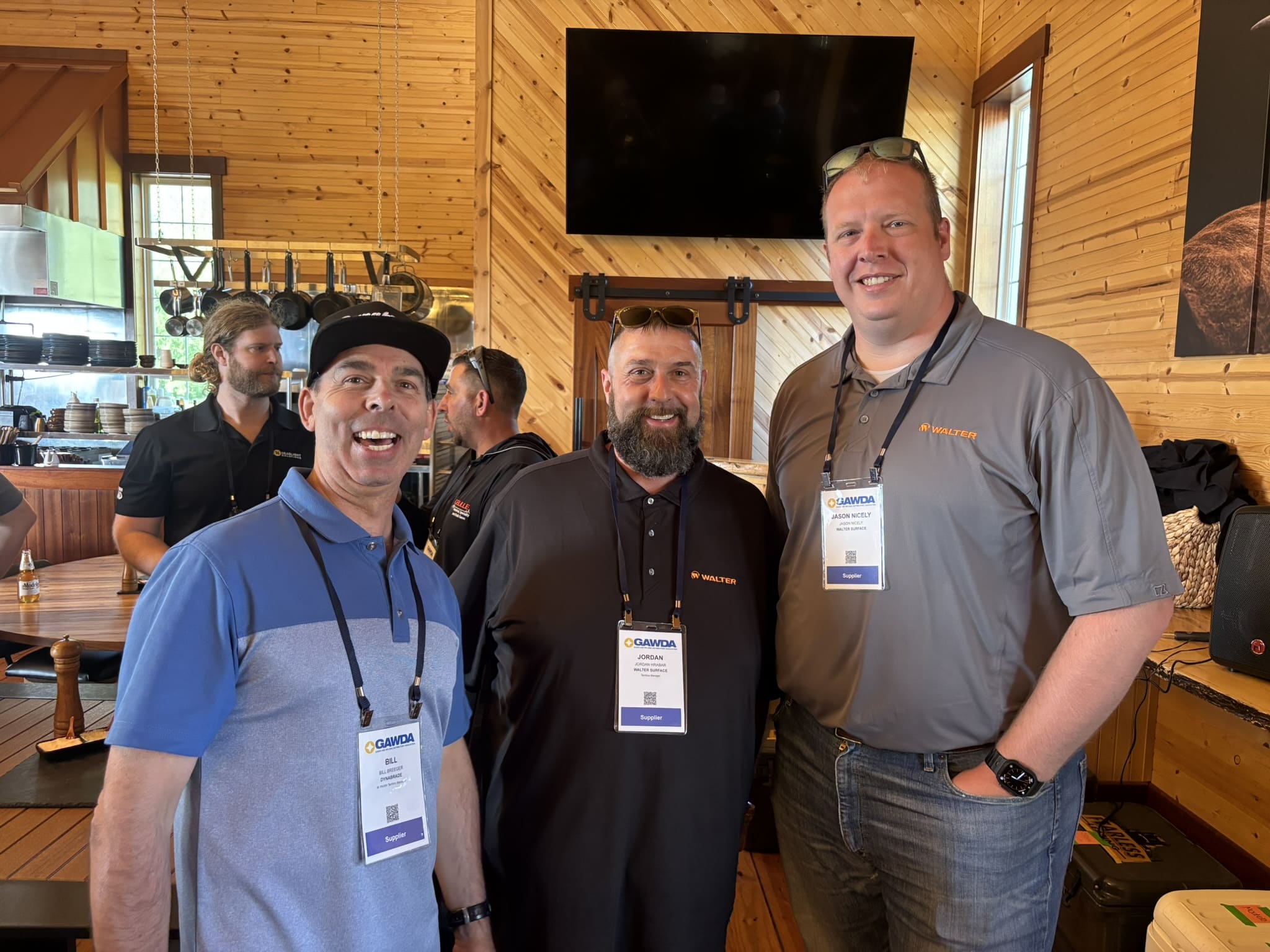 Four men wearing event badges pose for a group photo in a wood-paneled room, all smiling at the camera.