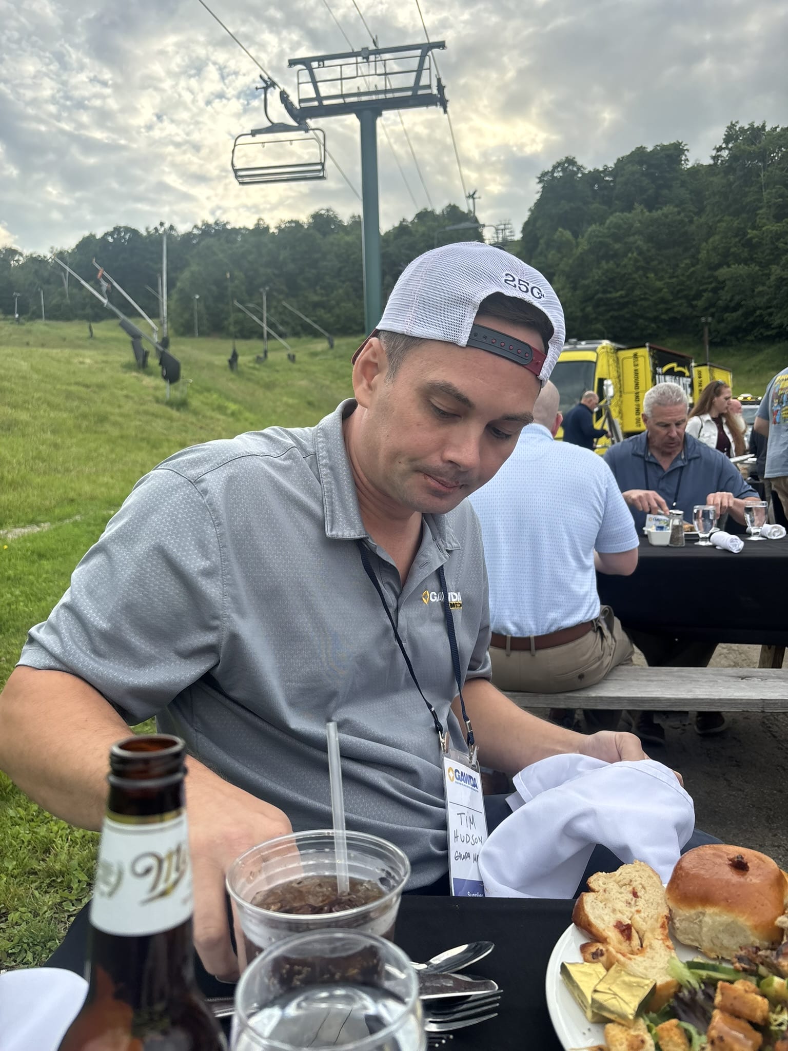 Man in a gray shirt and white cap sits at an outdoor picnic table with drinks and a plate of bread and salad; a ski lift and trees are in the background.