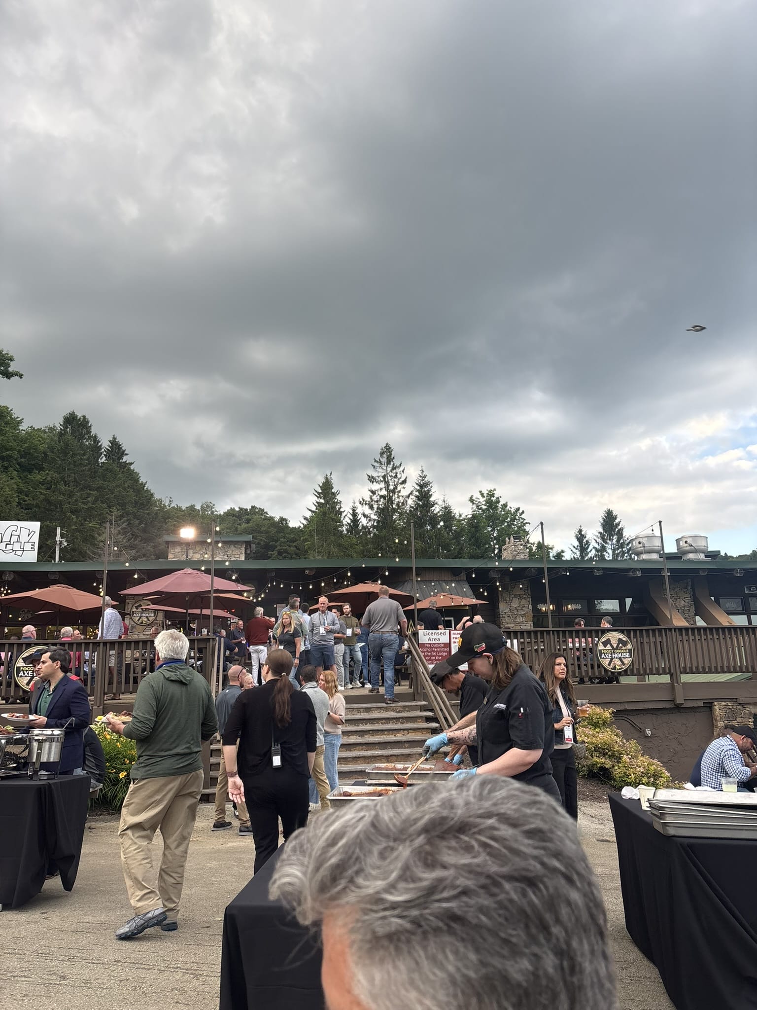 Crowd of people at an outdoor BBQ event under a cloudy sky, with grills and string lights overhead.