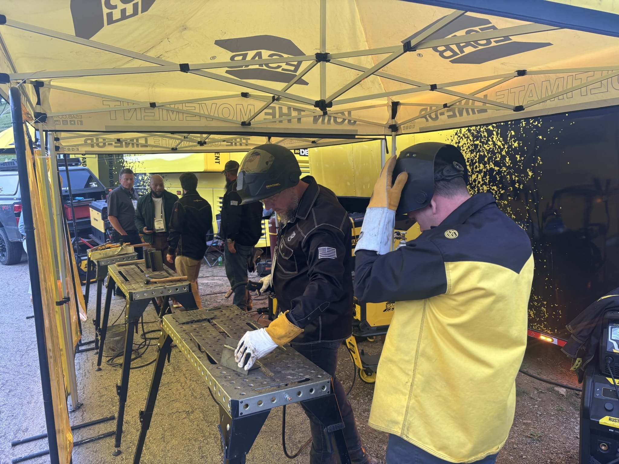 Group of workers under a yellow canopy, wearing helmets and gloves, standing around metal workbenches with tools.