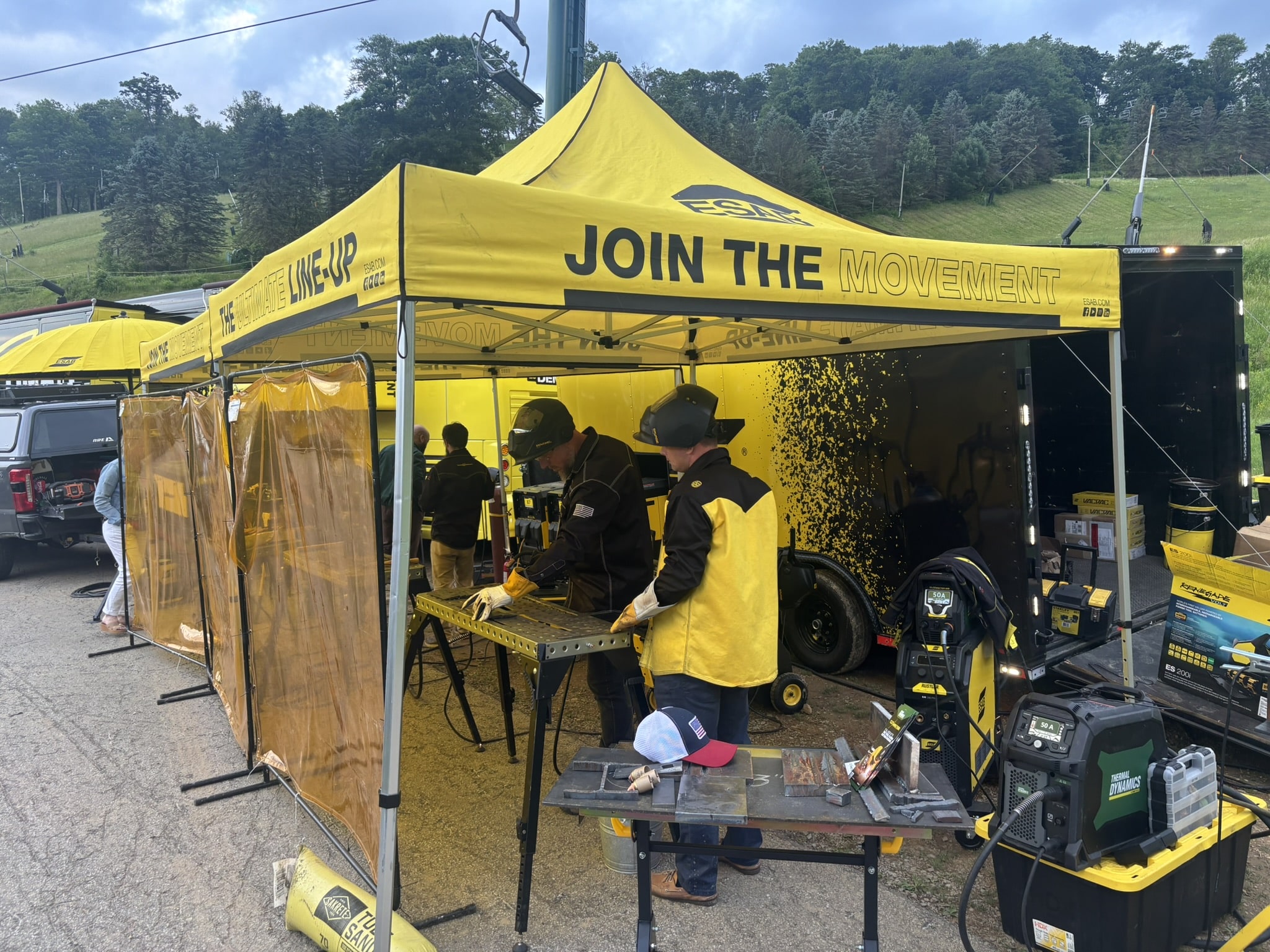 Welding demonstration booth under a bright yellow tent with 'JOIN THE MOVEMENT'; workers in protective gear weld at a workbench.