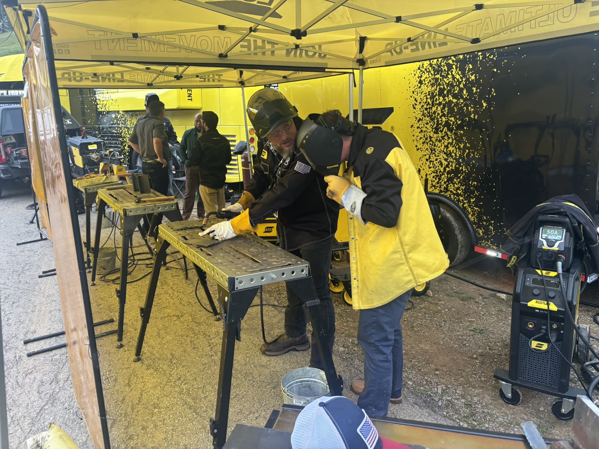 Welding workshop under a yellow canopy with workers in protective gear at a metal workbench.