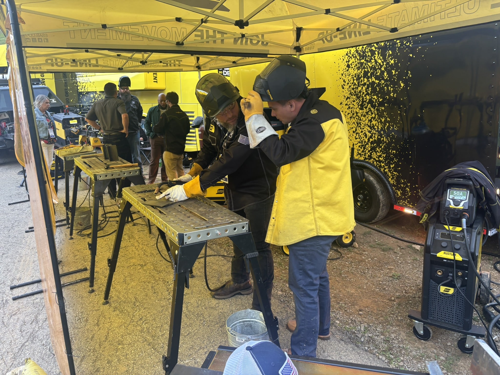 Group of people under a yellow canopy at a metal workshop; two workers in protective gear weld at a work table while others watch.