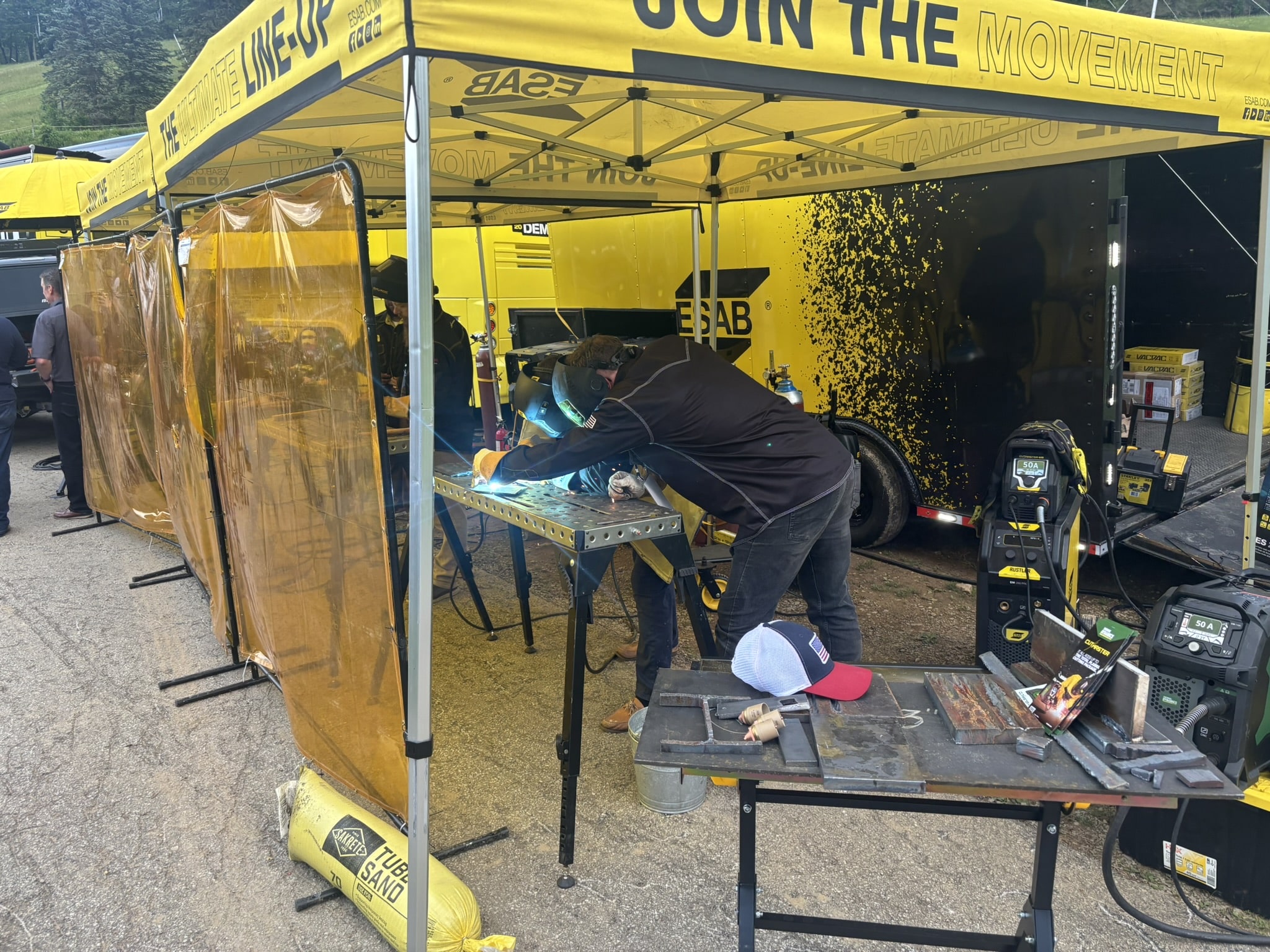 Welding work under a yellow tent with protective screens, a welder in dark clothing sparks at a metal table.