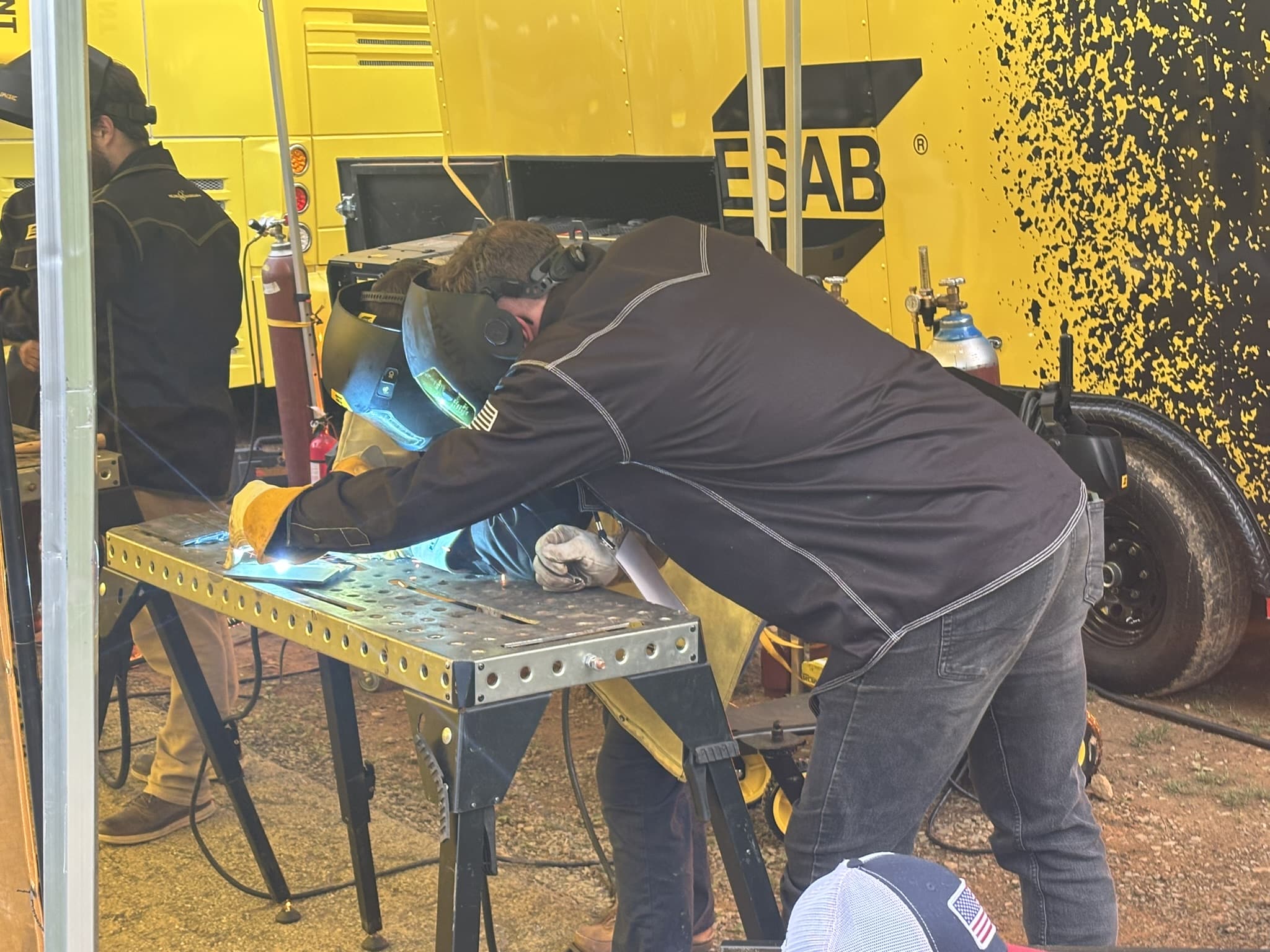 Welding worker wearing a helmet and protective gear leaning over a perforated metal workbench inside a yellow industrial trailer with tools and gas cylinders nearby