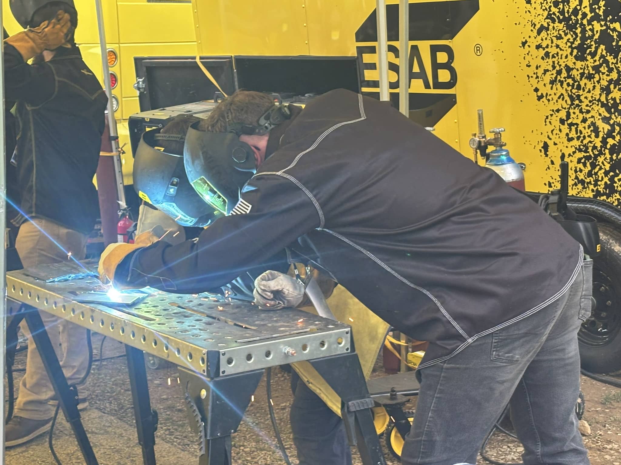 Welders in protective gear weld metal on a workbench, sparks flying, yellow vehicle in the background marked ESAB.