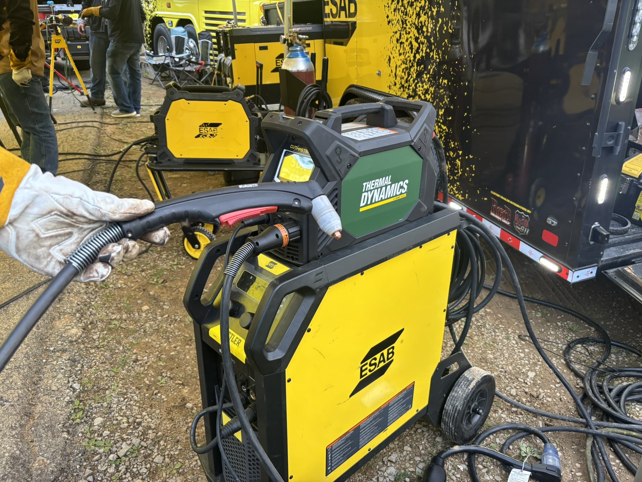 Industrial welding setup with yellow ESAB machines, hoses and cables on a gravel surface; workers and equipment in the background.