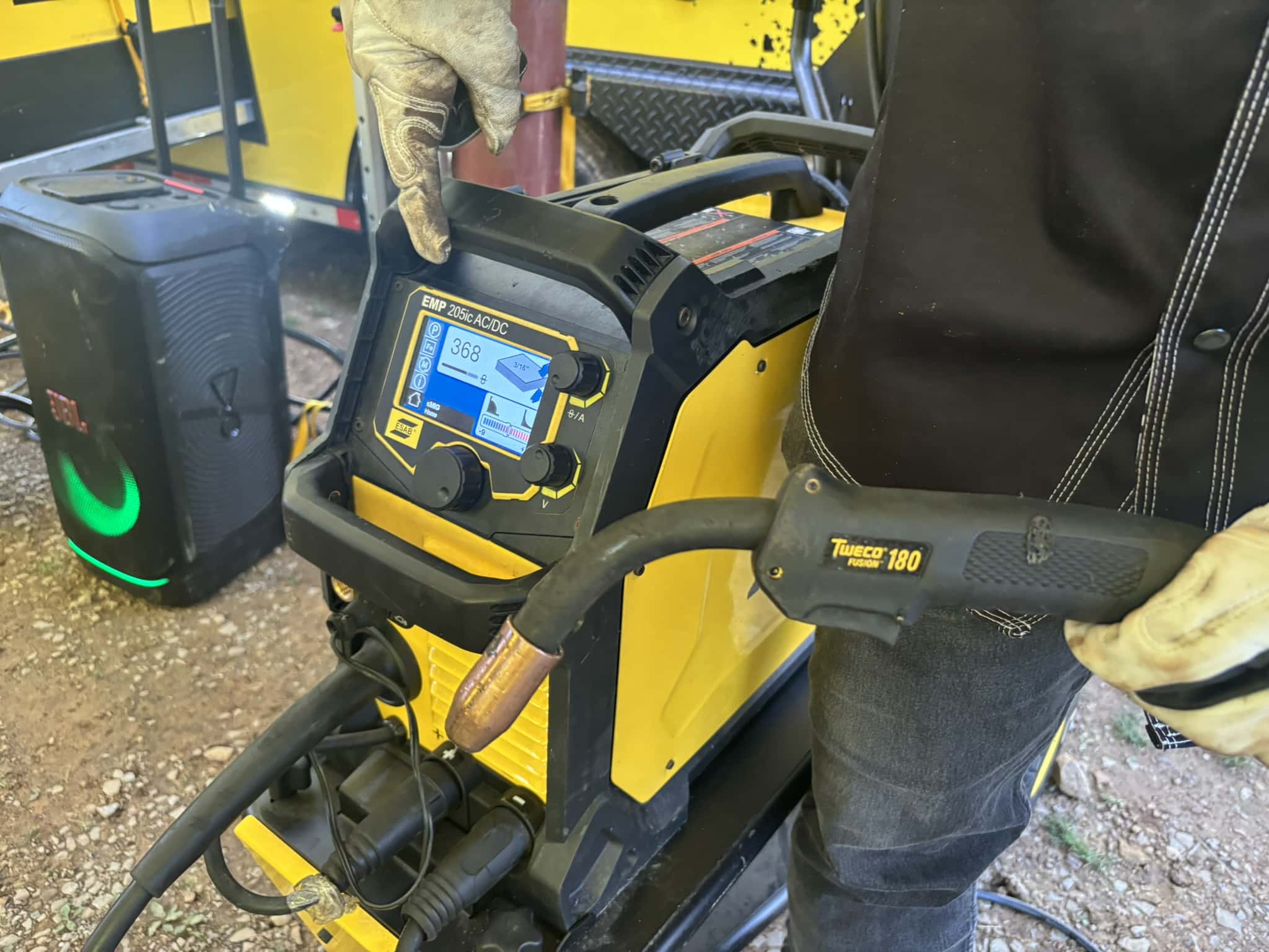 Gloved hand gripping the handle of a yellow welding machine with a digital display and controls visible on the front panel (Tweco Fusion 180).