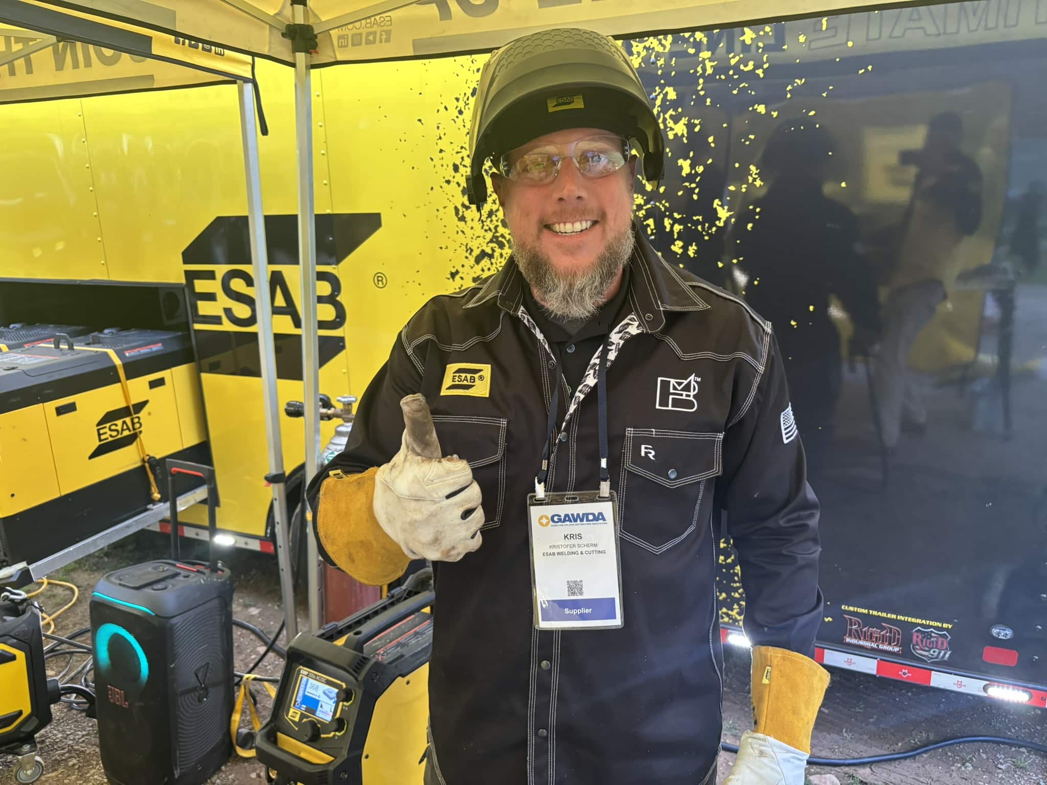 Welder wearing helmet and gloves, standing inside a yellow ESAB trailer with welding equipment nearby, badge visible at chest level.