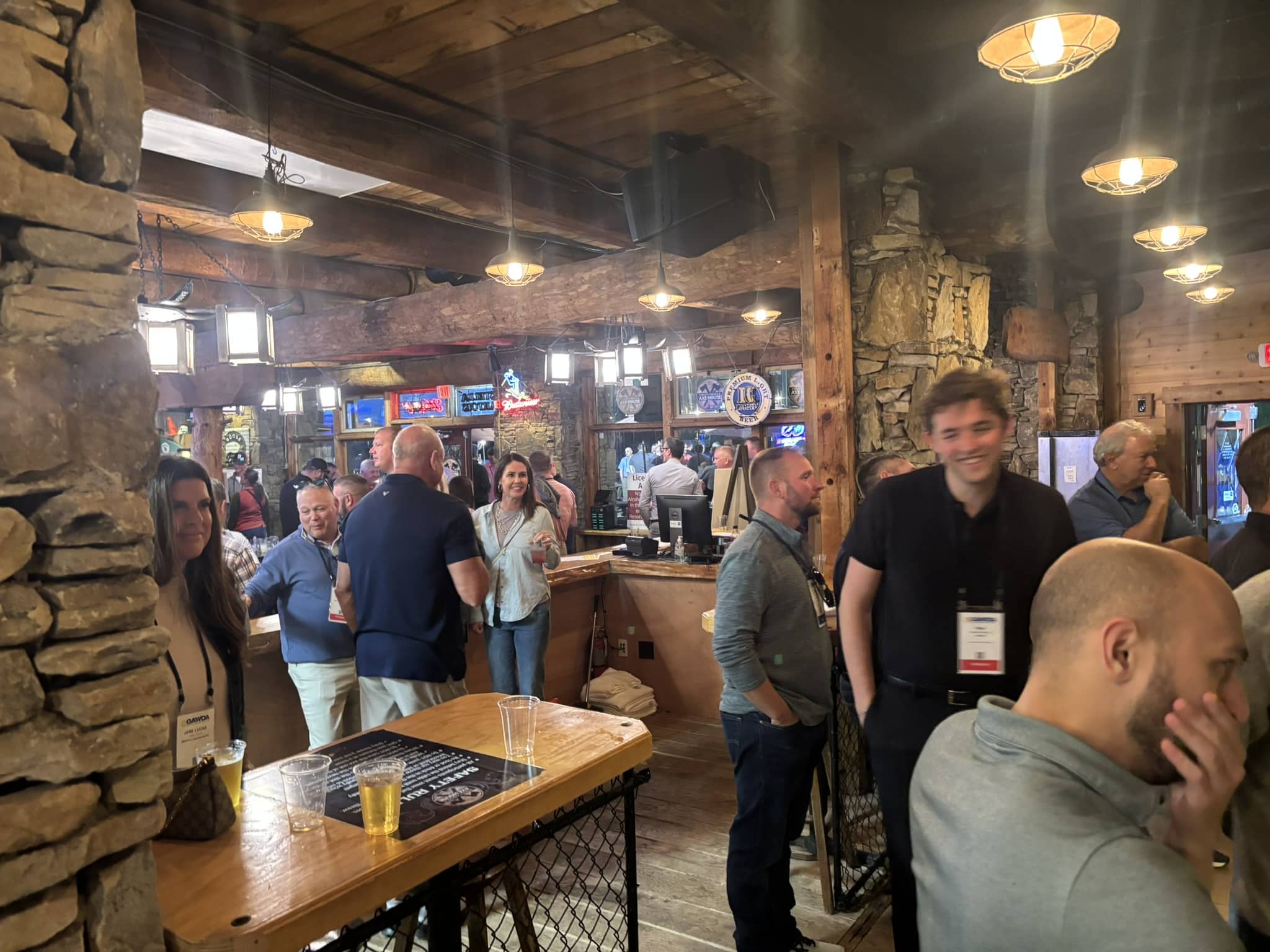 Bustling rustic bar interior with stone walls, wooden beams, and patrons standing at a long counter.