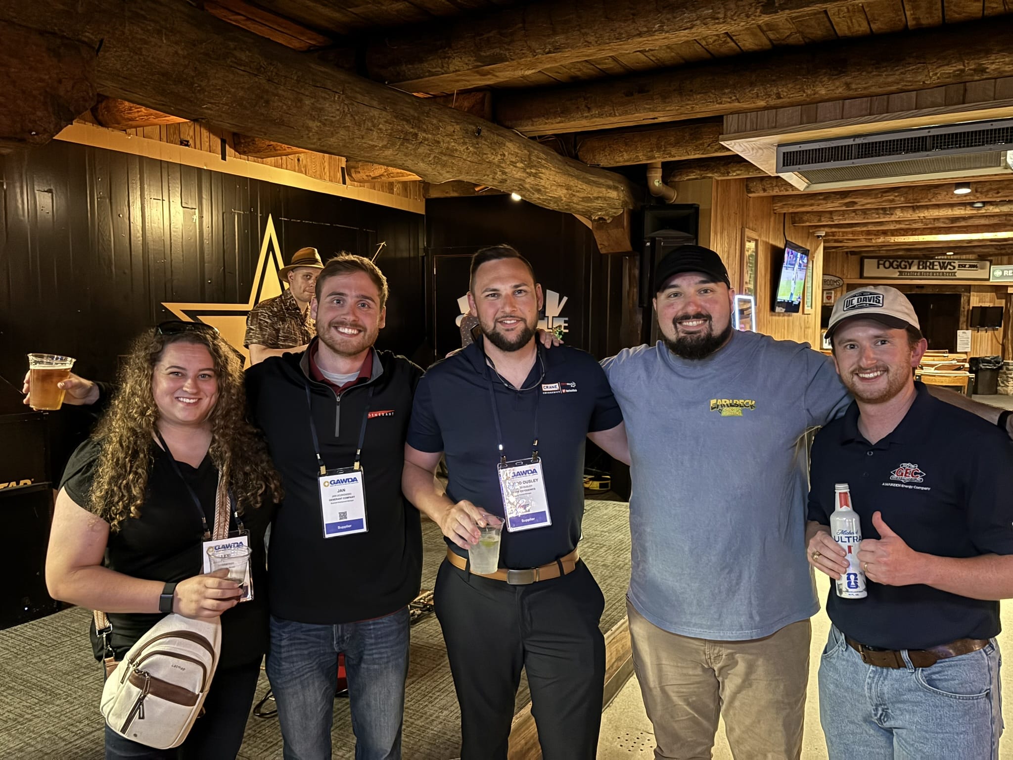Five adults posing together in a rustic bar area, smiling and holding drinks, wearing event badges around their necks.