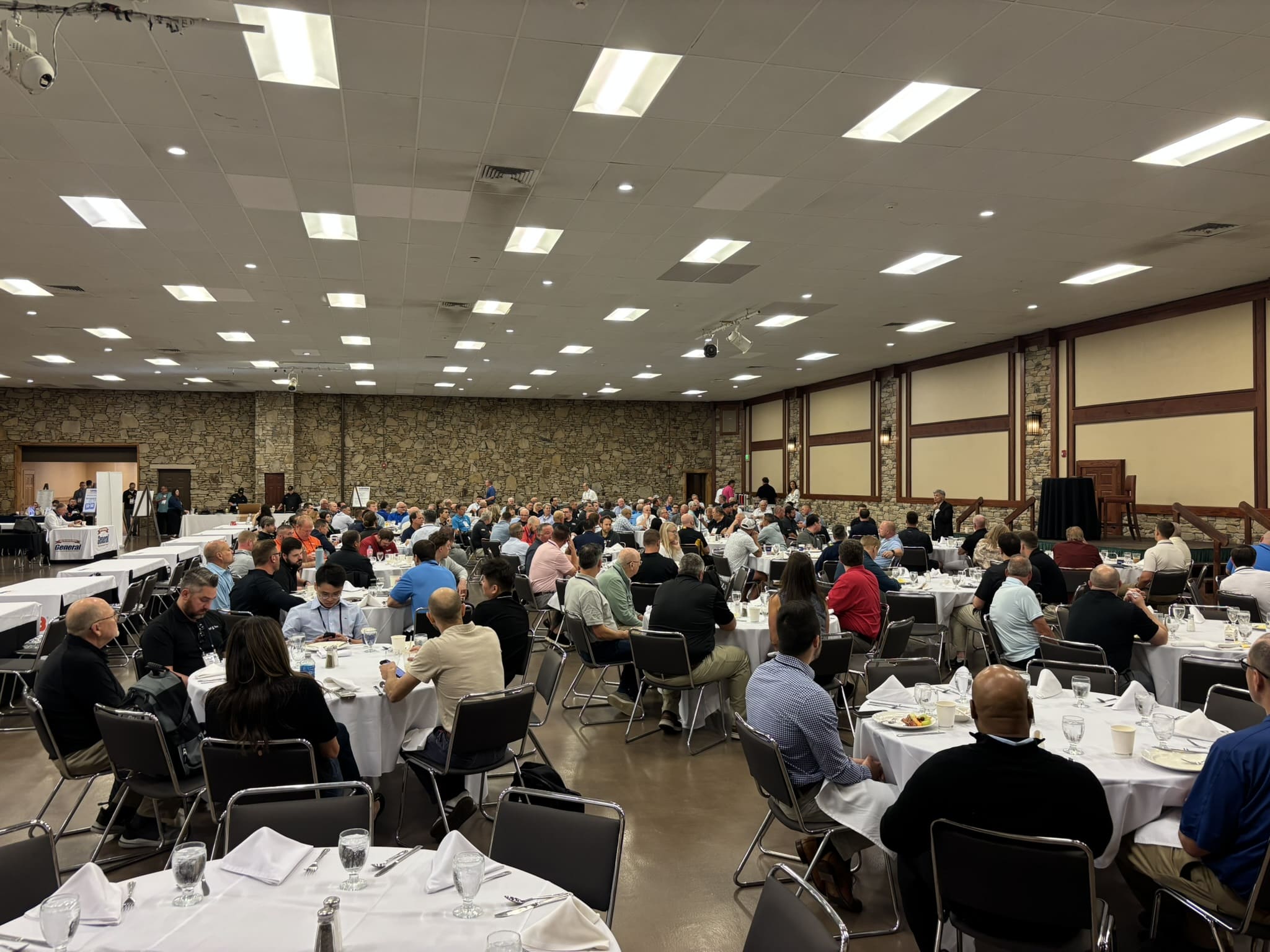 Large banquet hall filled with round tables of diners, white tablecloths, and glassware. Numerous attendees eat and chat in a stone-walled room.