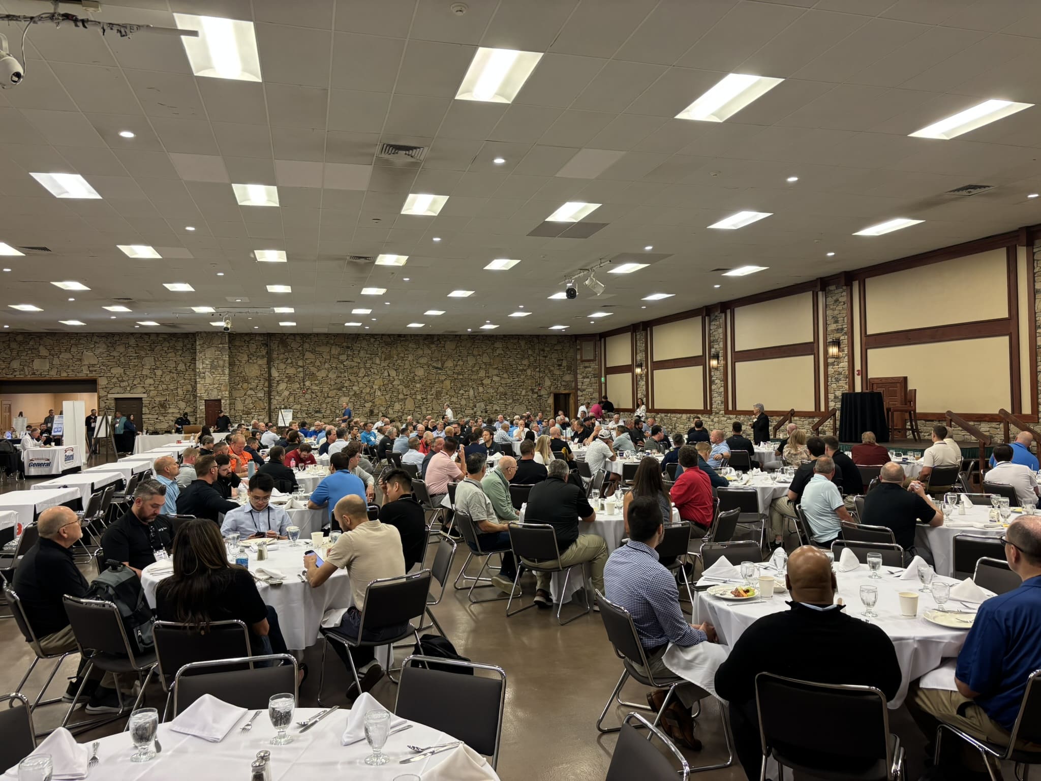 Banquet hall filled with round tables and dozens of attendees dining, with stone walls and bright ceiling lights.