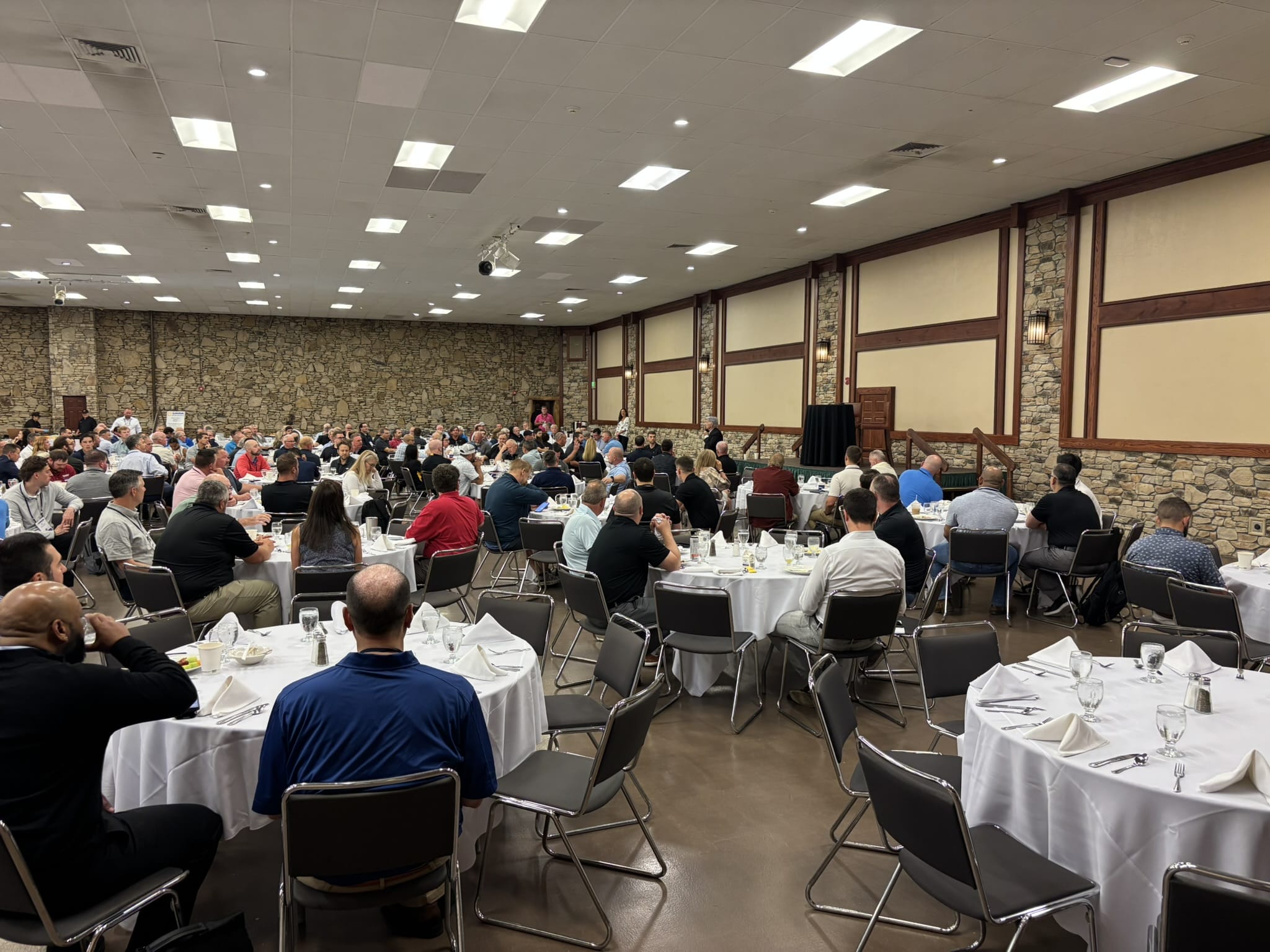 Large banquet hall filled with round tables and dozens of people dining and conversing, during a formal event notably with white tablecloths and glassware on tables.