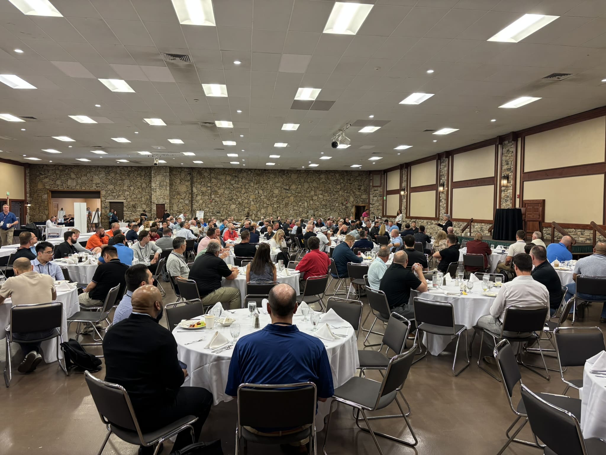Large banquet hall filled with people seated at round tables with white tablecloths for a conference or meal banquet.