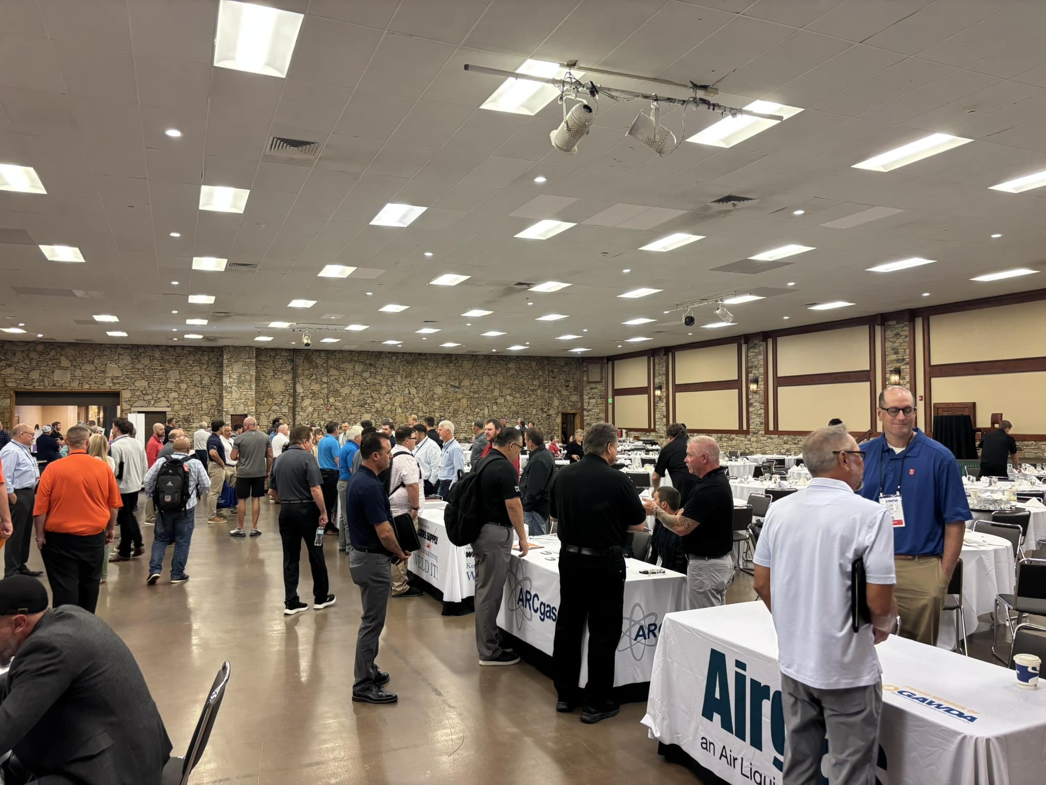 Crowd of people at a large indoor conference or trade show with display tables and banners nearby.