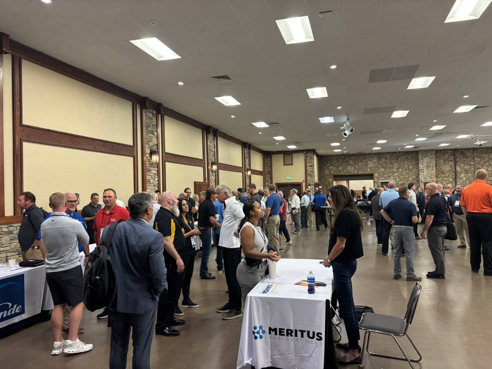 Crowded conference hall with attendees and exhibitor tables along stone walls, Meritus Gas Partners banners visible in the foreground.