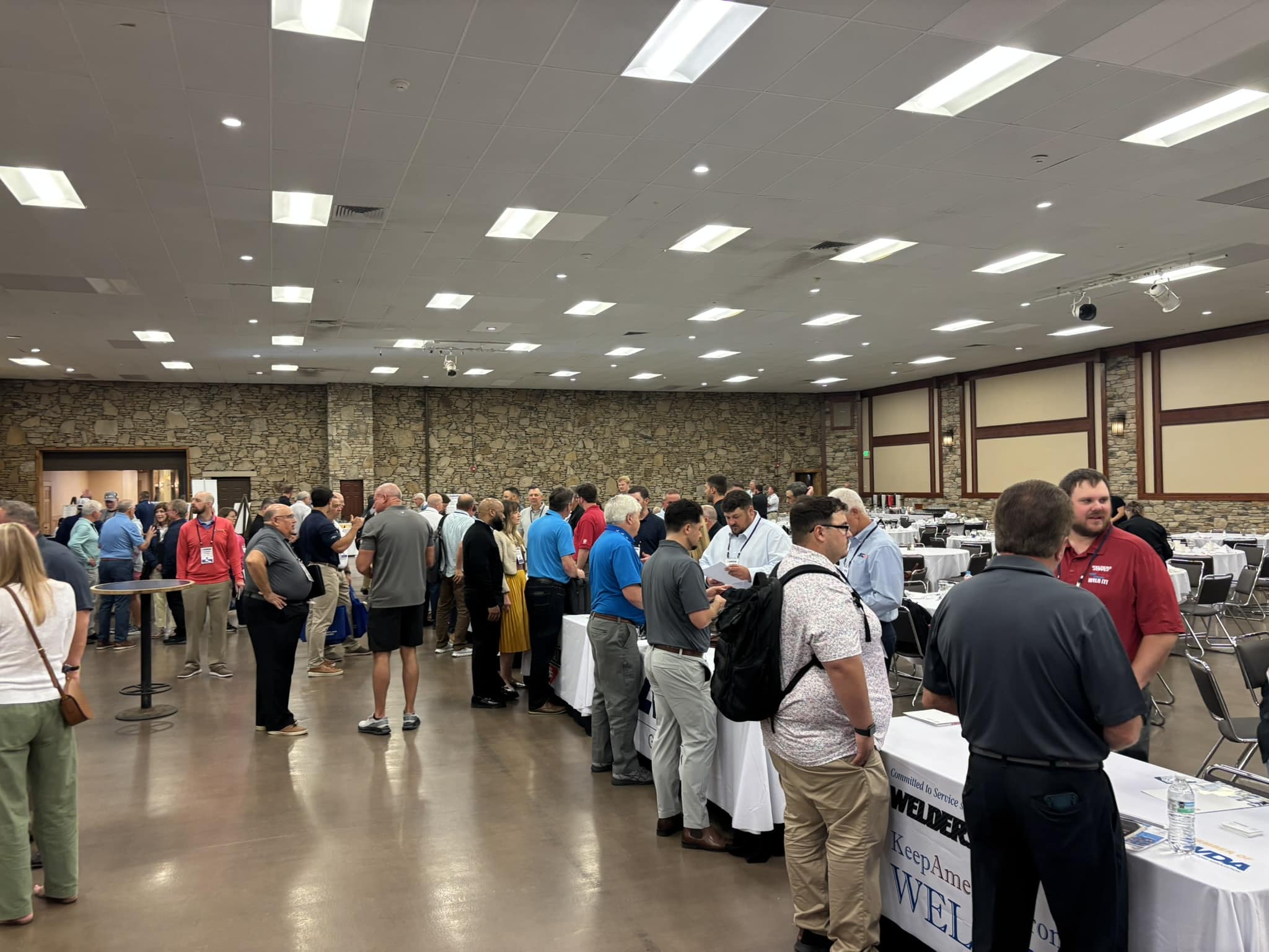 Busy conference hall filled with people standing around vendor tables and banners along a stone wall.