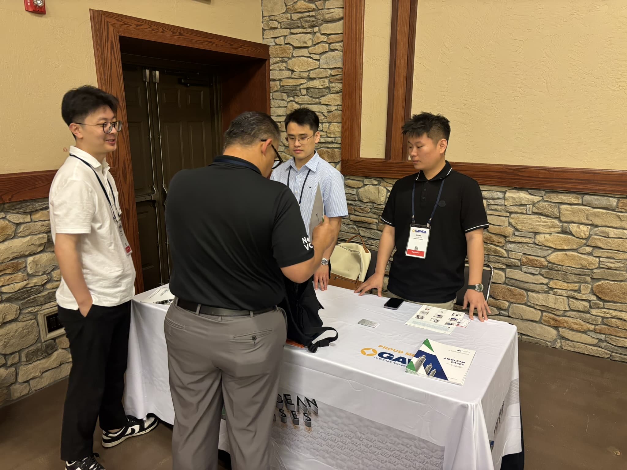 Group of men in business casual gathered around a table with brochures and name badges, discussing documents in a hallway with stone walls and wood trim.