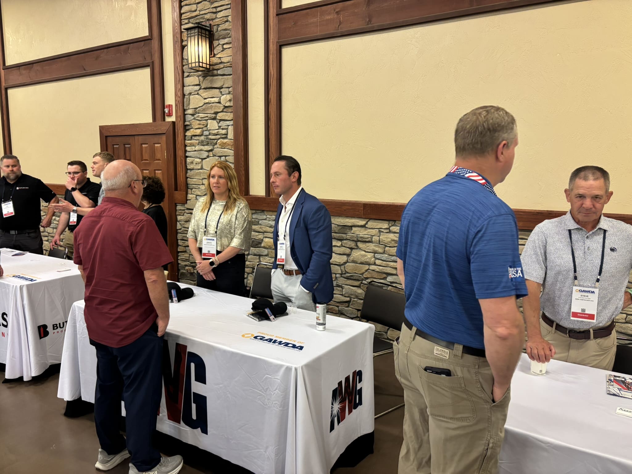 Group of conference attendees standing at a booth with white tablecloths and GAWDA logos, in an indoor event space with stone accents and warm lighting.